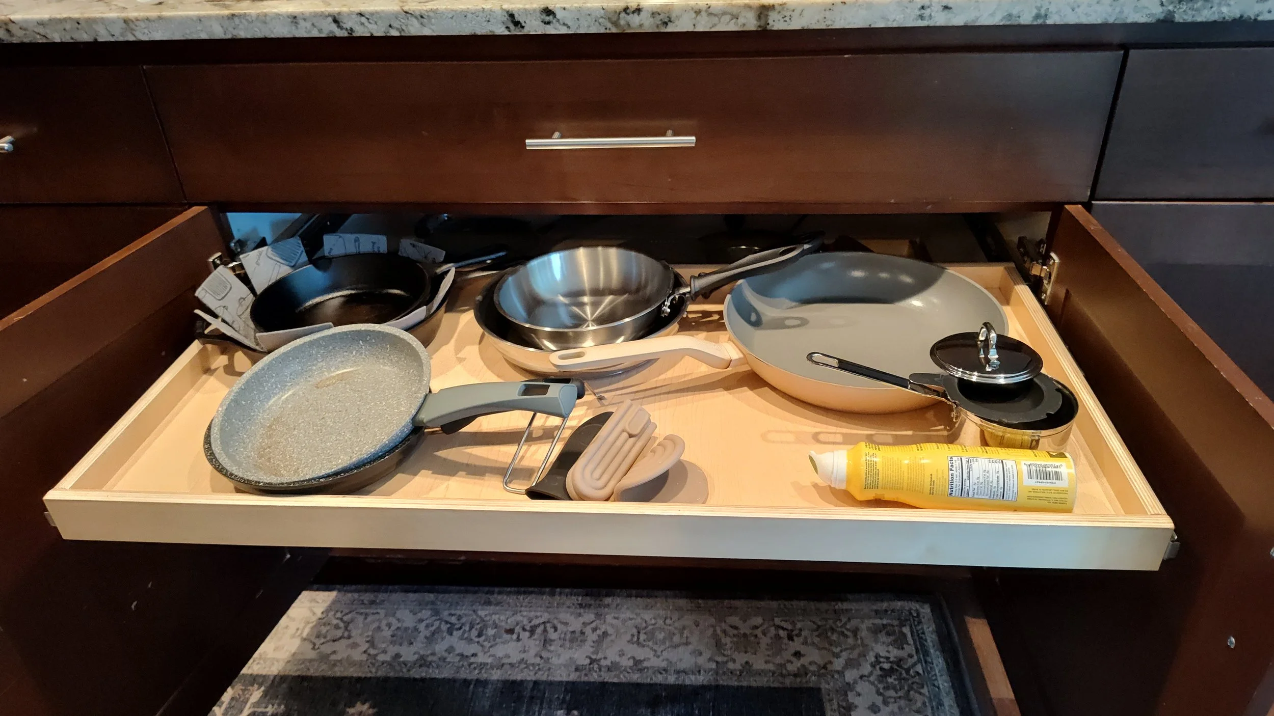 Kitchen drawer with various frying pans, a grater, rubber spatula, and a bottle of cooking spray.
