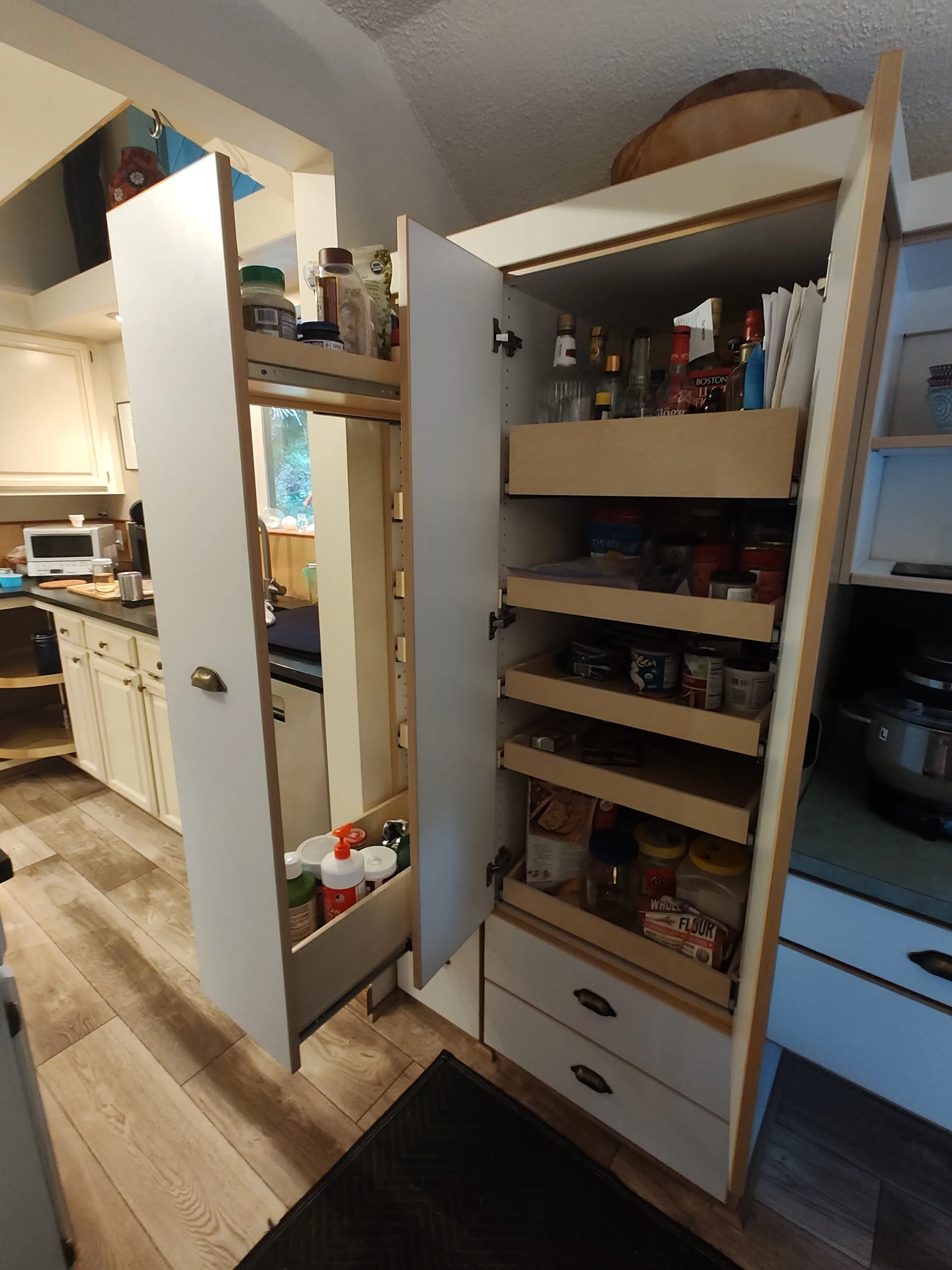 Open kitchen pantry with snacks, jars, spices, and cooking supplies on shelves, next to a kitchen counter with appliances and utensils.