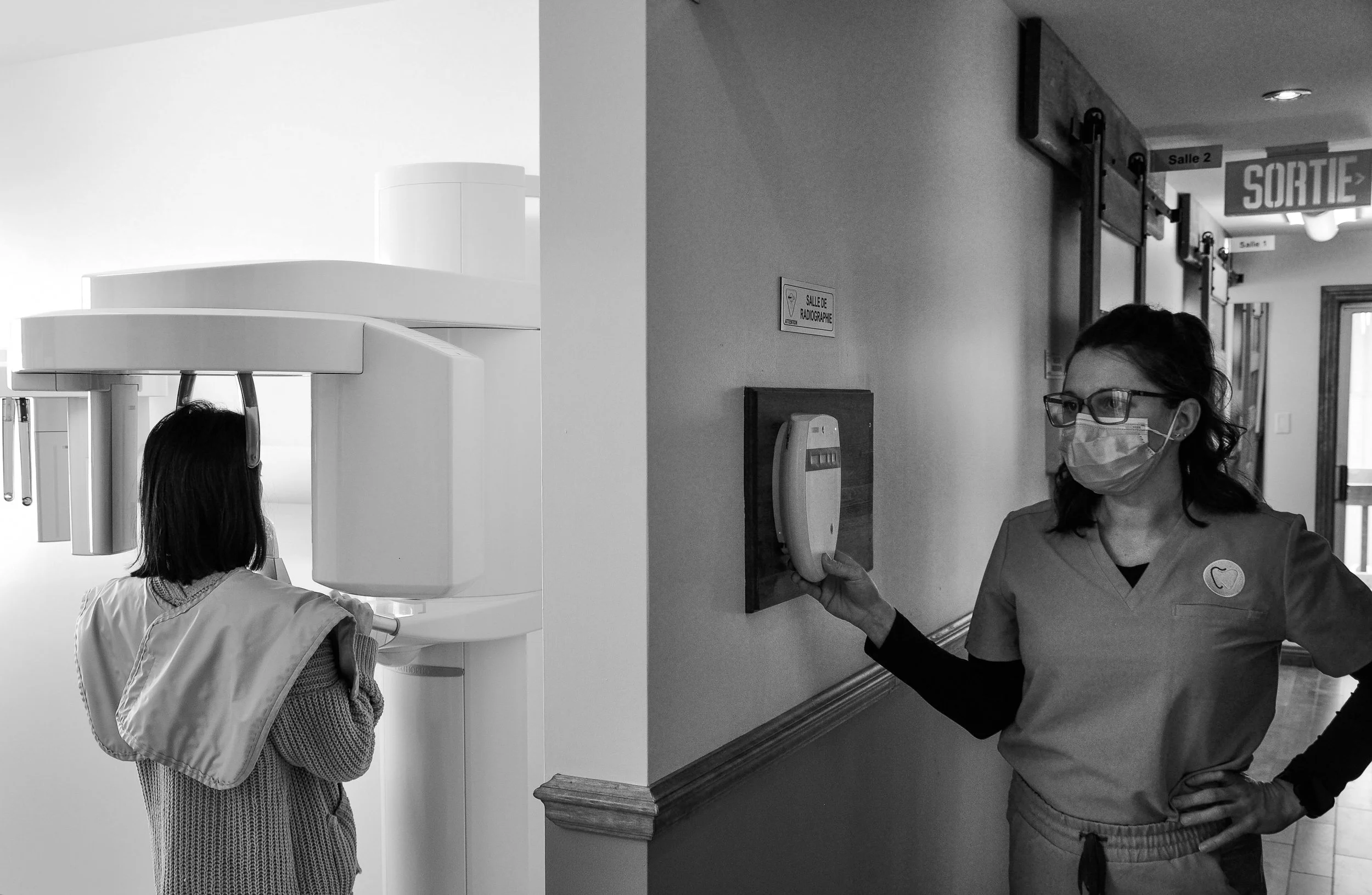Vue en noir et blanc d'une assistante dentaire opérant une machine de radiographie panoramique dentaire pendant qu'une patiente passe son examen dans une salle dédiée.