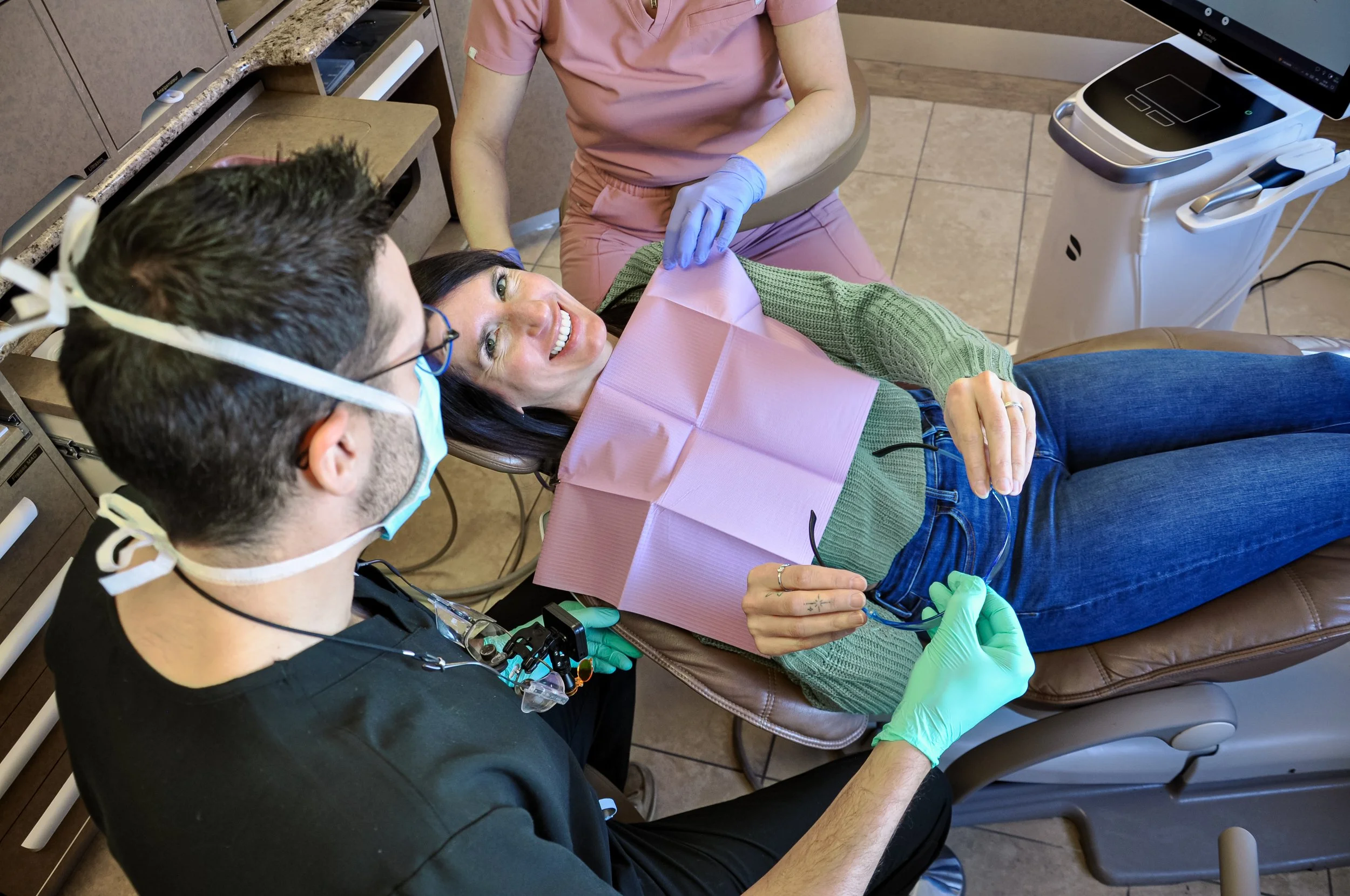 Une patiente souriante allongée sur un fauteuil de dentiste, entourée d'un dentiste et d'une assistante préparant une consultation dentaire avec un équipement moderne en arrière-plan.