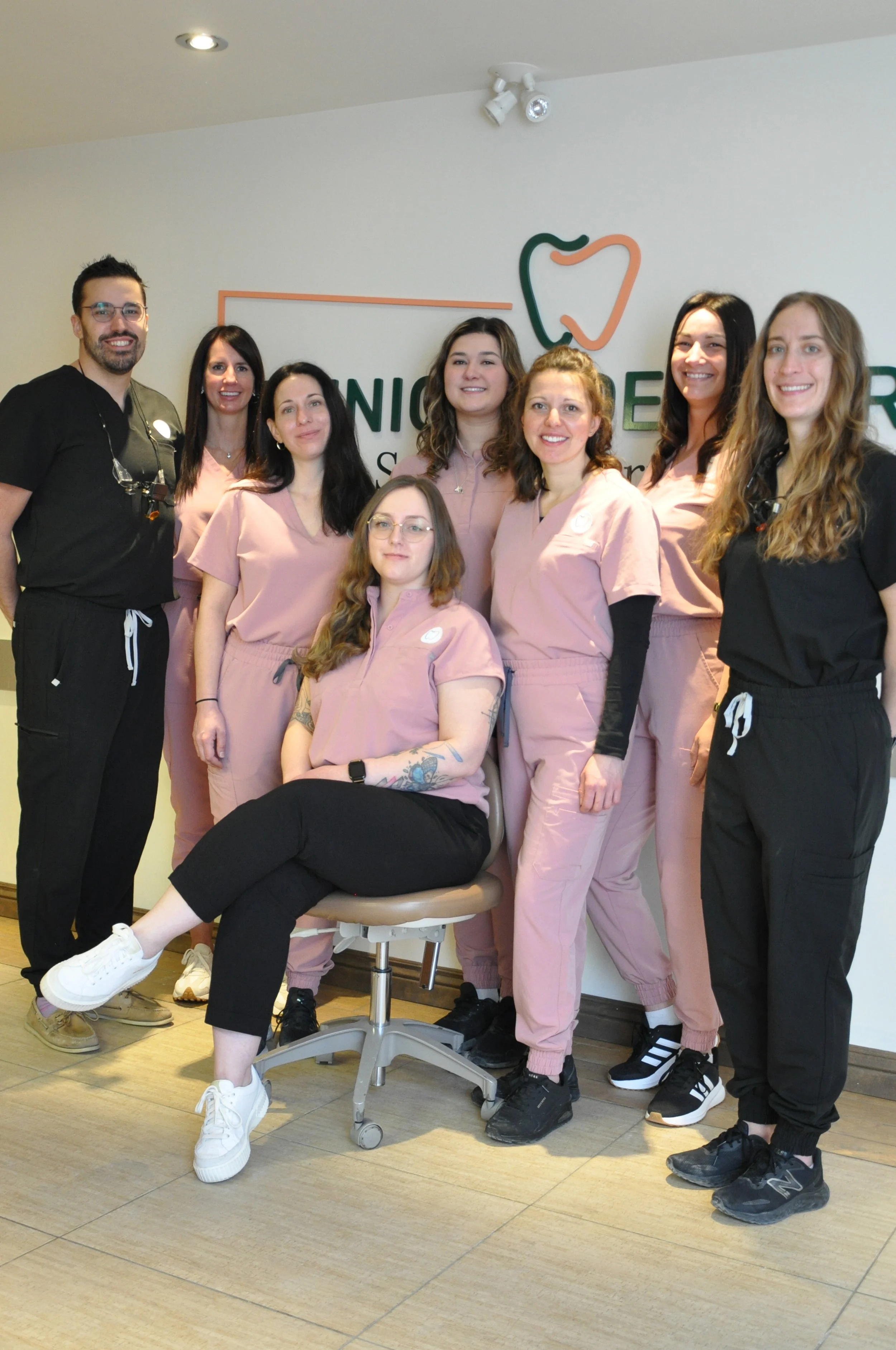 Portrait de l'équipe de la Clinique Dentaire St-Élie d'Orford, composée de dentistes et d'assistantes souriants en uniformes cliniques roses et noirs, posant devant le logo de la clinique.