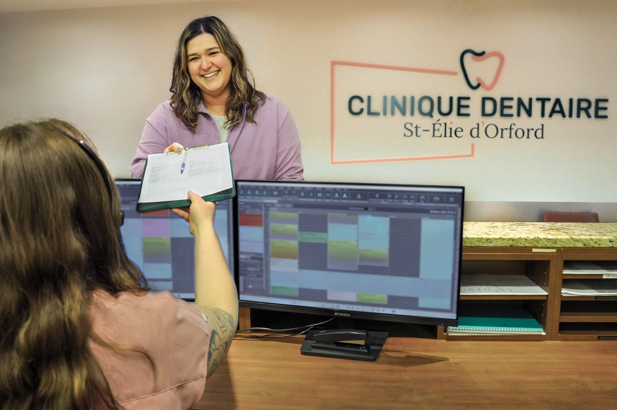 Une patiente souriante à la réception de la Clinique Dentaire St-Élie d'Orford remet un formulaire sur un porte-bloc à la secrétaire médicale devant son écran d'ordinateur."