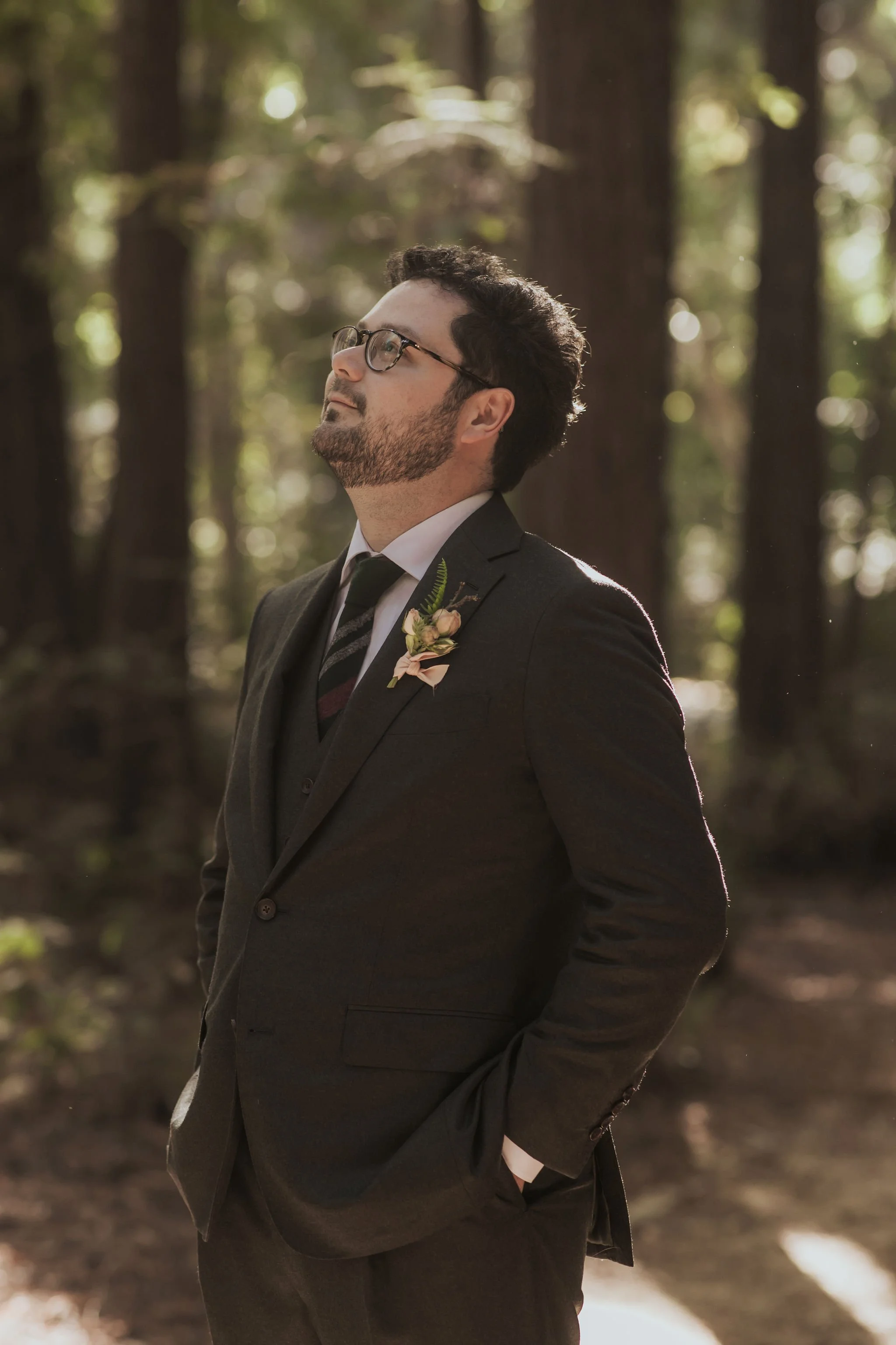 A man in a black suit with a boutonniere, glasses, and dark hair standing outdoors in a forest, looking up in a moment of contemplation.