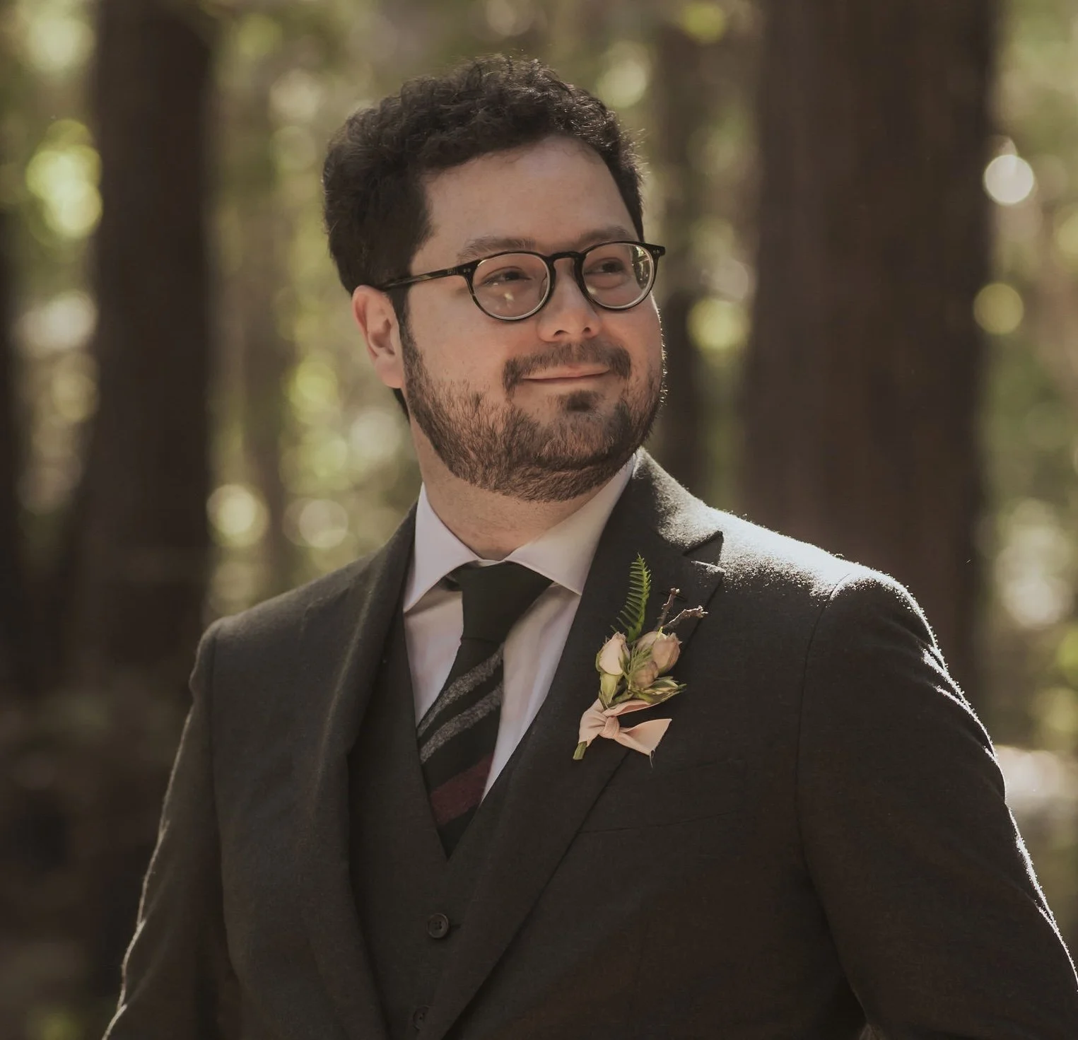 A man in a suit with glasses standing in a forest with sunlight shining through the trees.