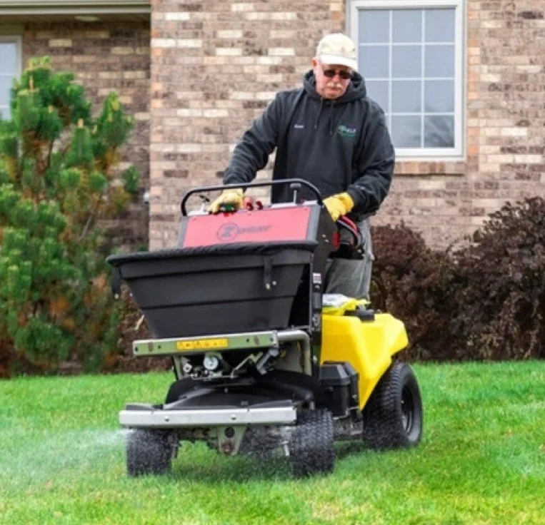 A man operating a yellow and black turf blower on a lawn in front of a brick house with a window and bushes.