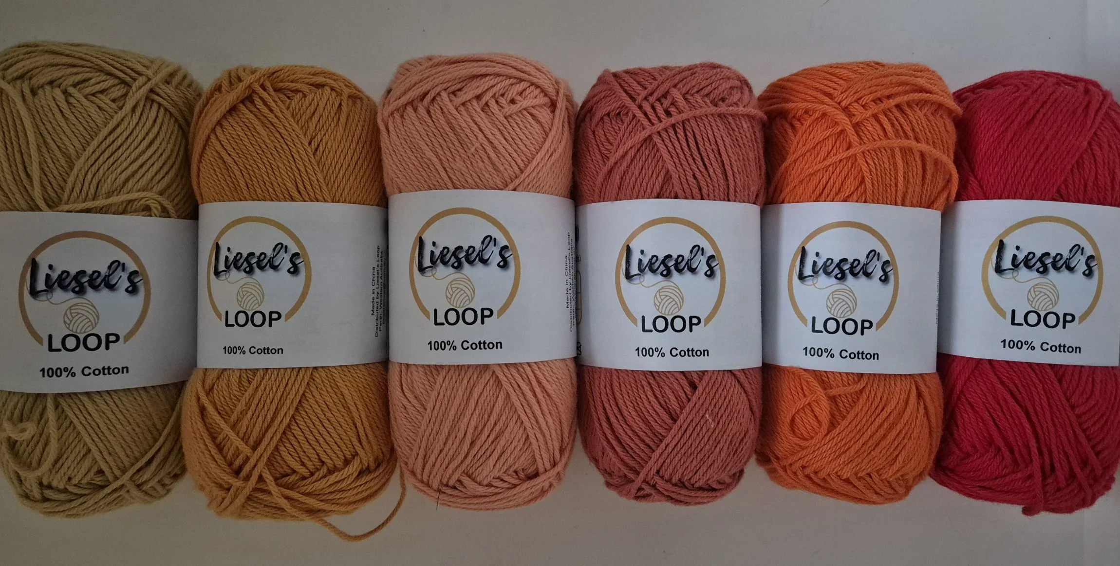 Six yarn skeins labeled 'Liesel's Loop,' made of 100% cotton, in shades of yellow, orange, pink, peach, and red, arranged in a row.