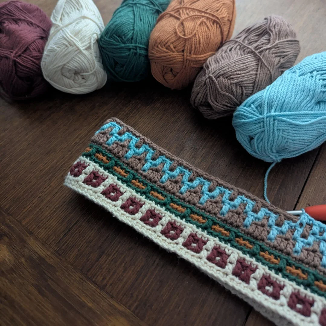 Assorted balls of yarn in colors including maroon, cream, green, tan, gray, and blue, with a partially completed crochet project featuring various colorful patterns lying on a wooden surface.