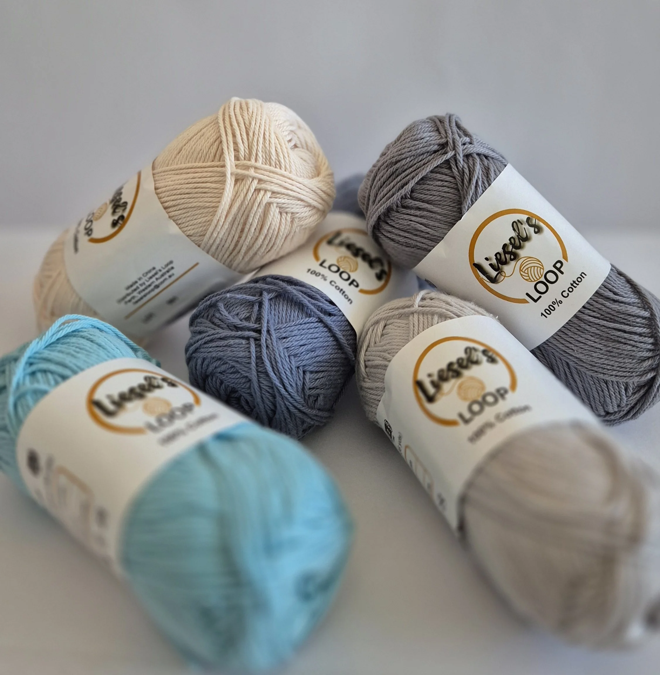 Close-up of balls of yarn in light cream, gray, and blue colors, with labels indicating they are 100% cotton, branded "Liese's Loop".
