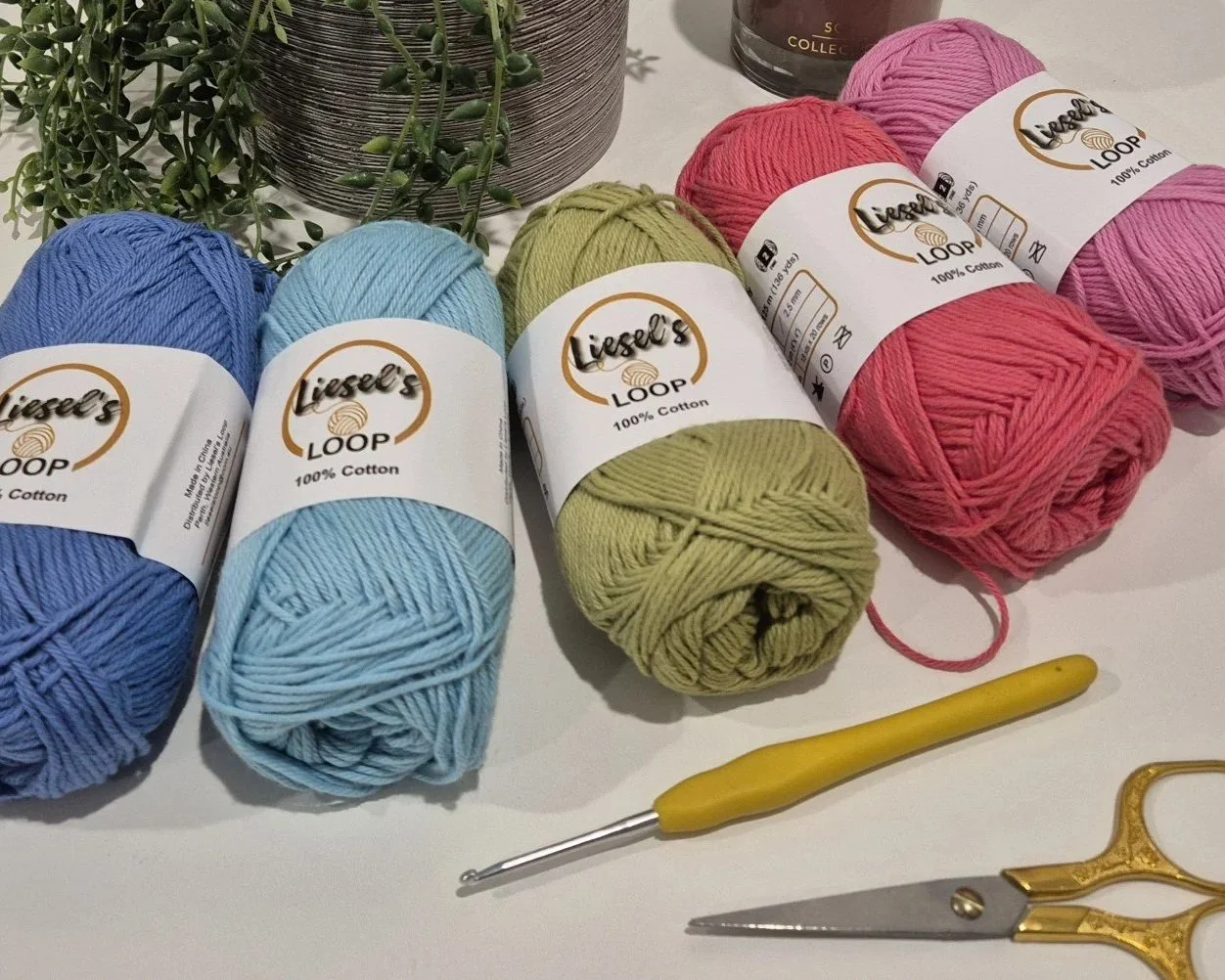 Six balls of colorful Liesel's Loop yarn in Blue, Light Blue, Artichoke, Bubblegum, and Candy Floss, with a small yellow crochet hook and gold scissors on a white surface. A potted green plant is partially visible in the background.