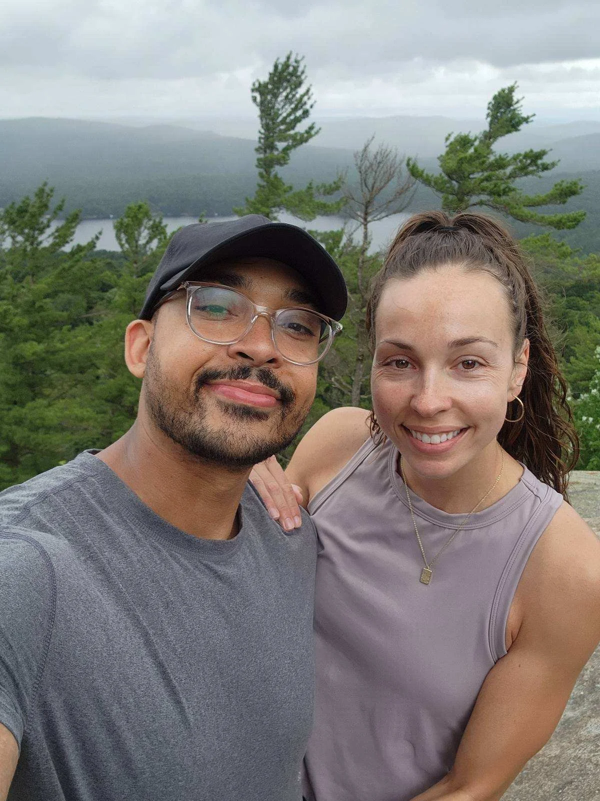 A smiling man and woman taking a selfie outdoors on a cloudy day with trees and a body of water in the background.