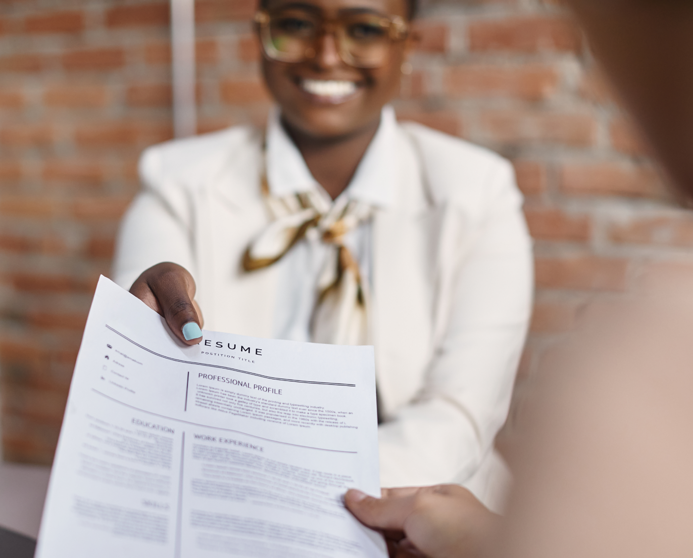 Smiling woman in glasses and white blazer handing a resume to someone.