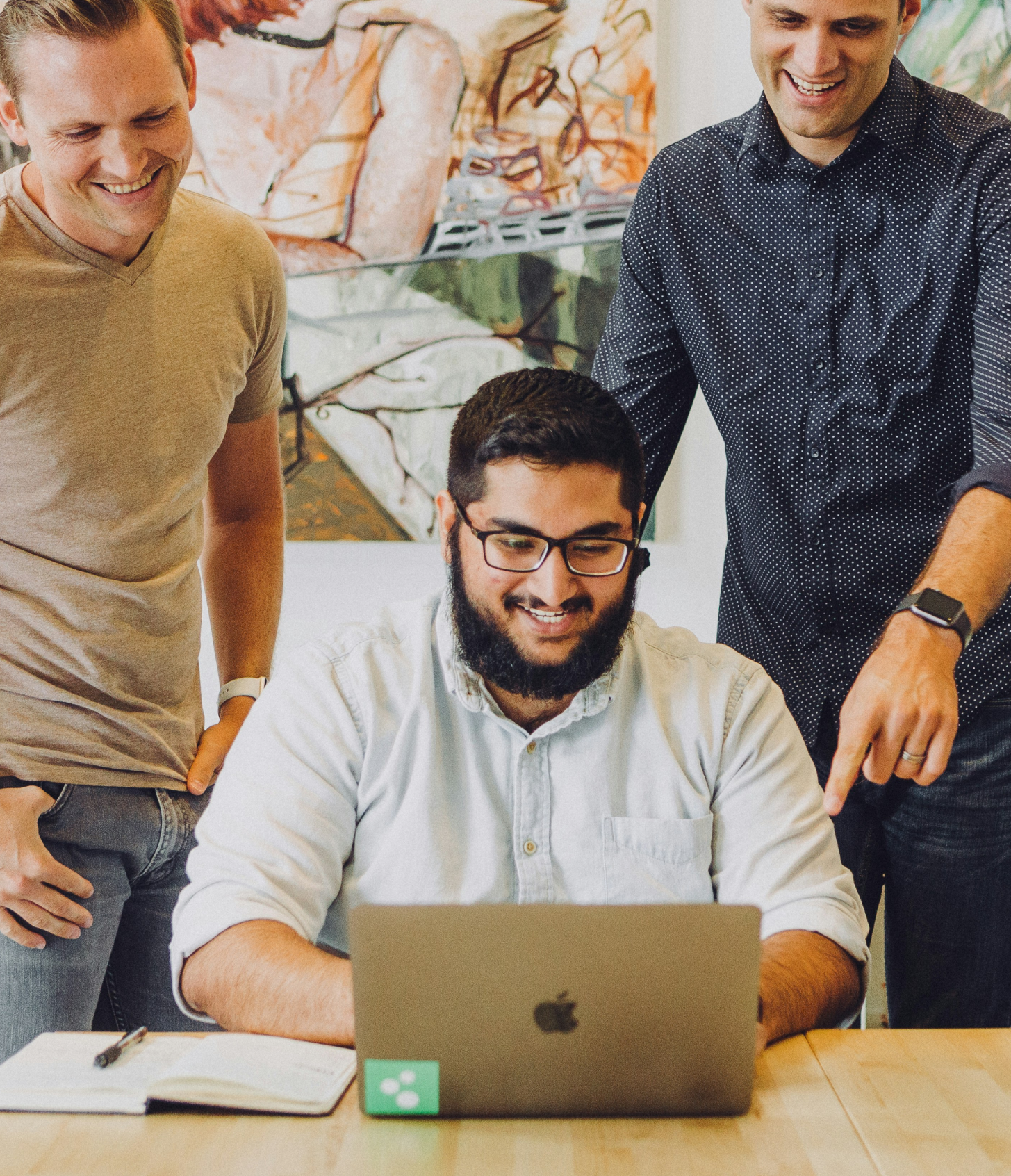 Three men are gathered around a man sitting at a desk, working on a laptop and smiling. The other two men are standing and leaning in, also smiling, in an office setting with artwork on the wall.