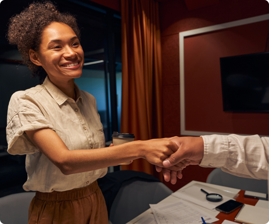 A woman smiling and shaking hands with another person in an office setting.