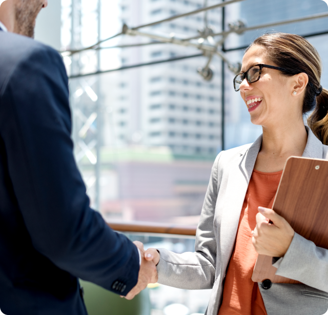 A woman with glasses, holding a clipboard, smiling, shaking hands with a man in a business suit inside a modern office building with large windows and cityscape view.