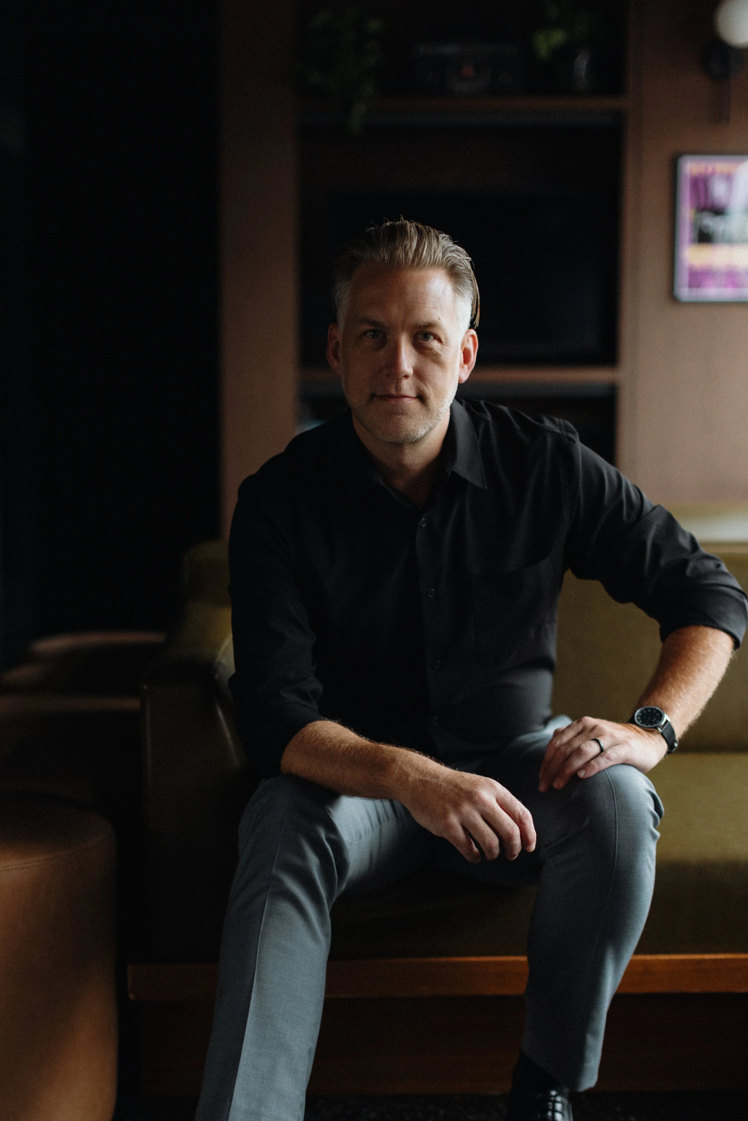 The author, a man with light hair, wearing a black shirt, seated on a green sofa in a dimly lit room, looking directly at the camera.