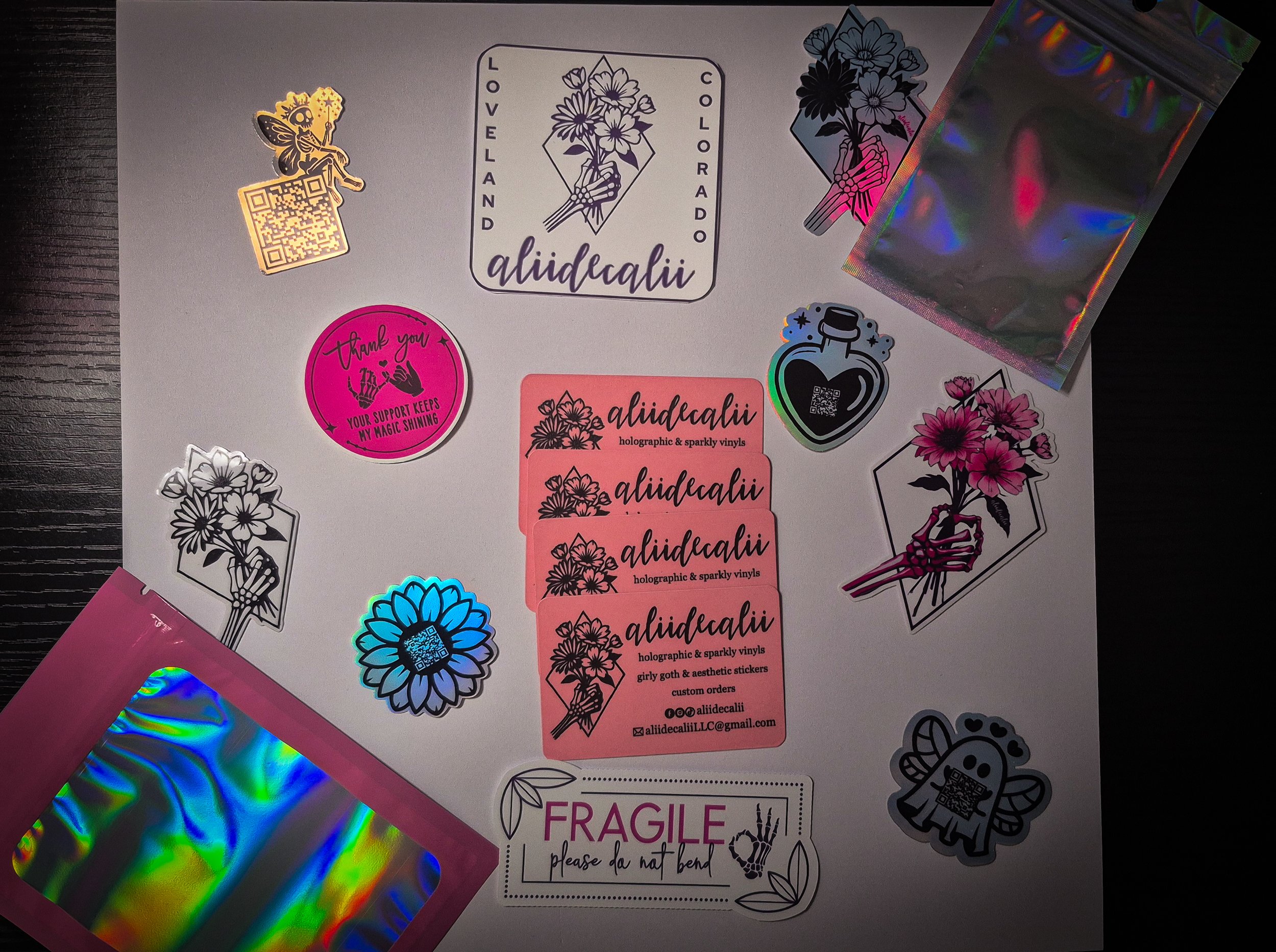 A sheet of decorative stickers featuring flowers, a fairy, and a girl skeleton, alongside holographic and sparkly vinyl sticker sheets, and a pink 'FRAGILE' warning sticker on a black surface.