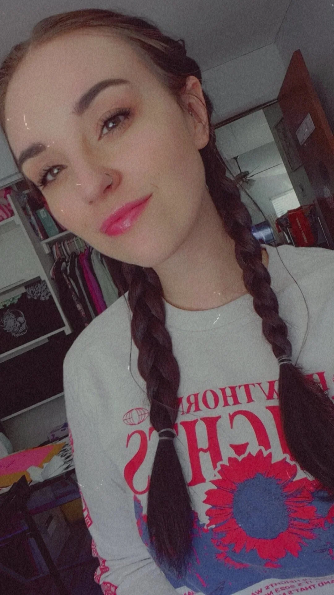A young woman with braided dark hair, fair skin, and makeup, wearing a gray shirt with red and blue floral graphics and text, taking a selfie in a room with clothing and shelving in the background.