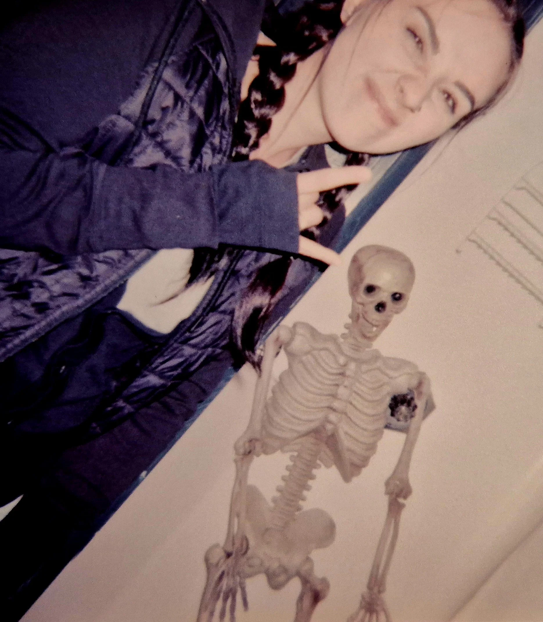 A person with long dark hair and a dark jacket making a hand gesture, next to a skeleton model in an educational setting.