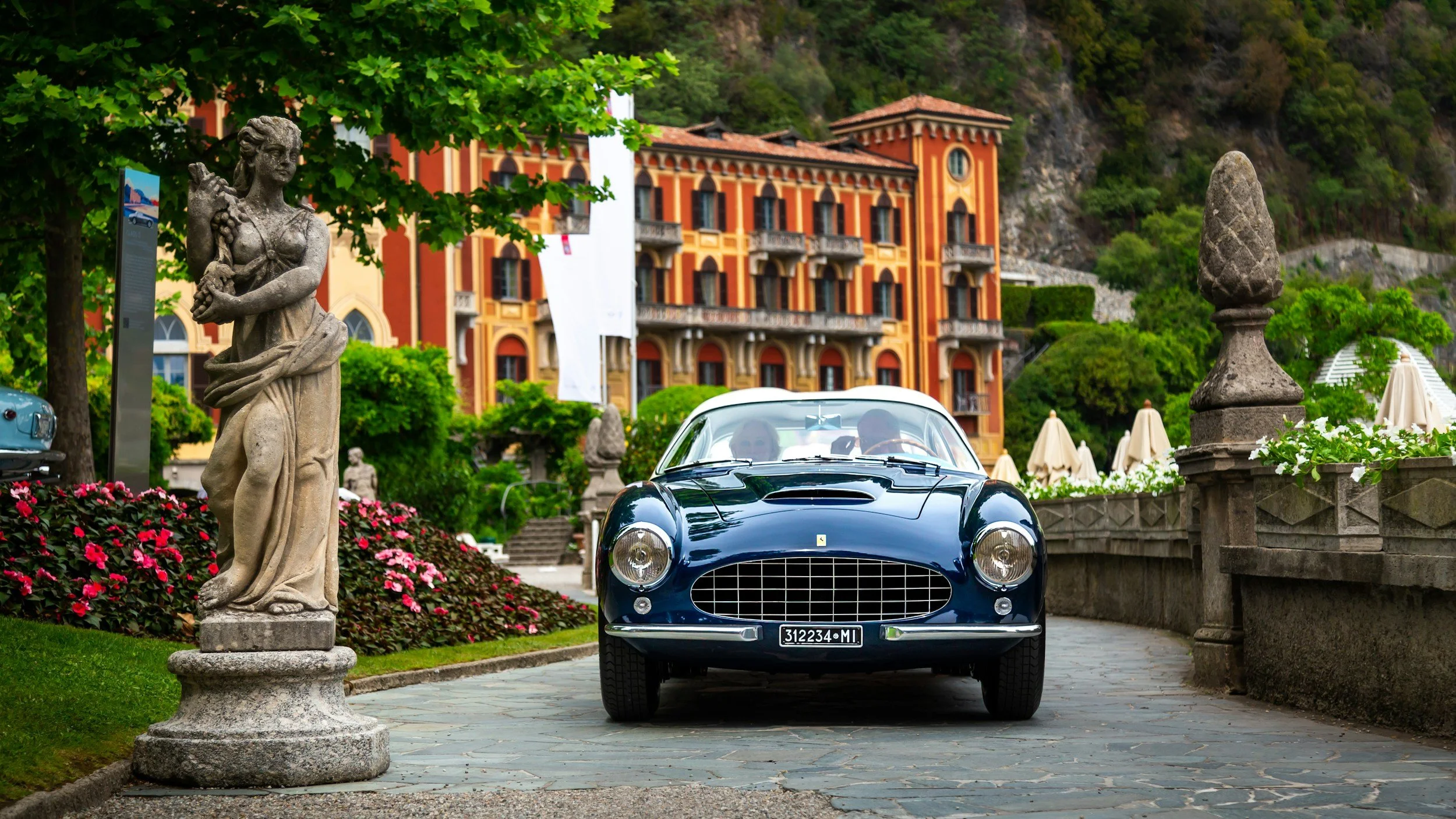 A vintage blue Ferrari parked on a cobblestone path surrounded by classical statues, lush greenery, and a grand building with orange and yellow accents in the background.