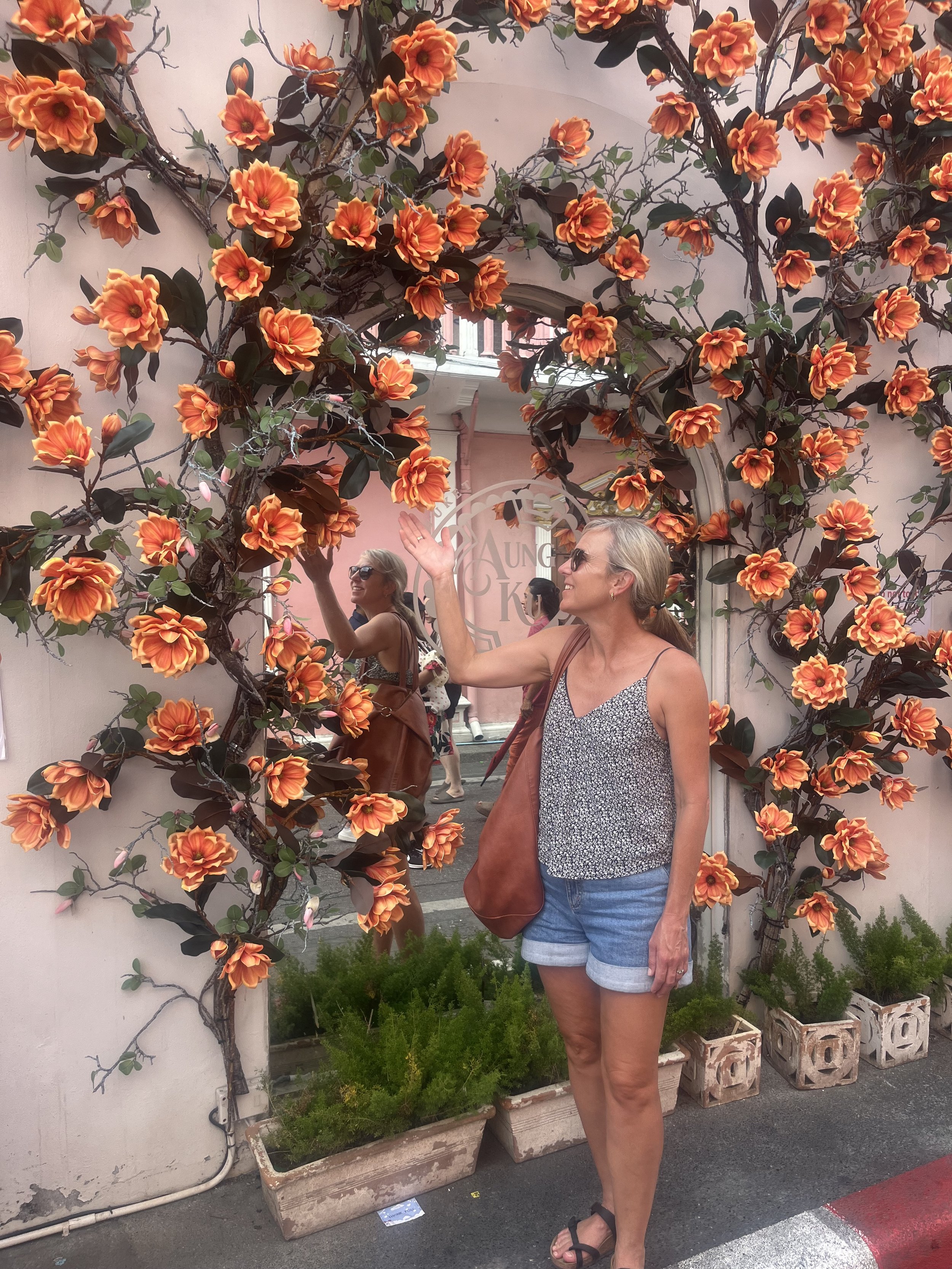 A woman wearing sunglasses, a sleeveless top, and denim shorts stands in front of a decorative pink wall with a mirror and orange flower vines. She is reaching toward the mirror with her right hand, and a reflection of herself can be seen.
