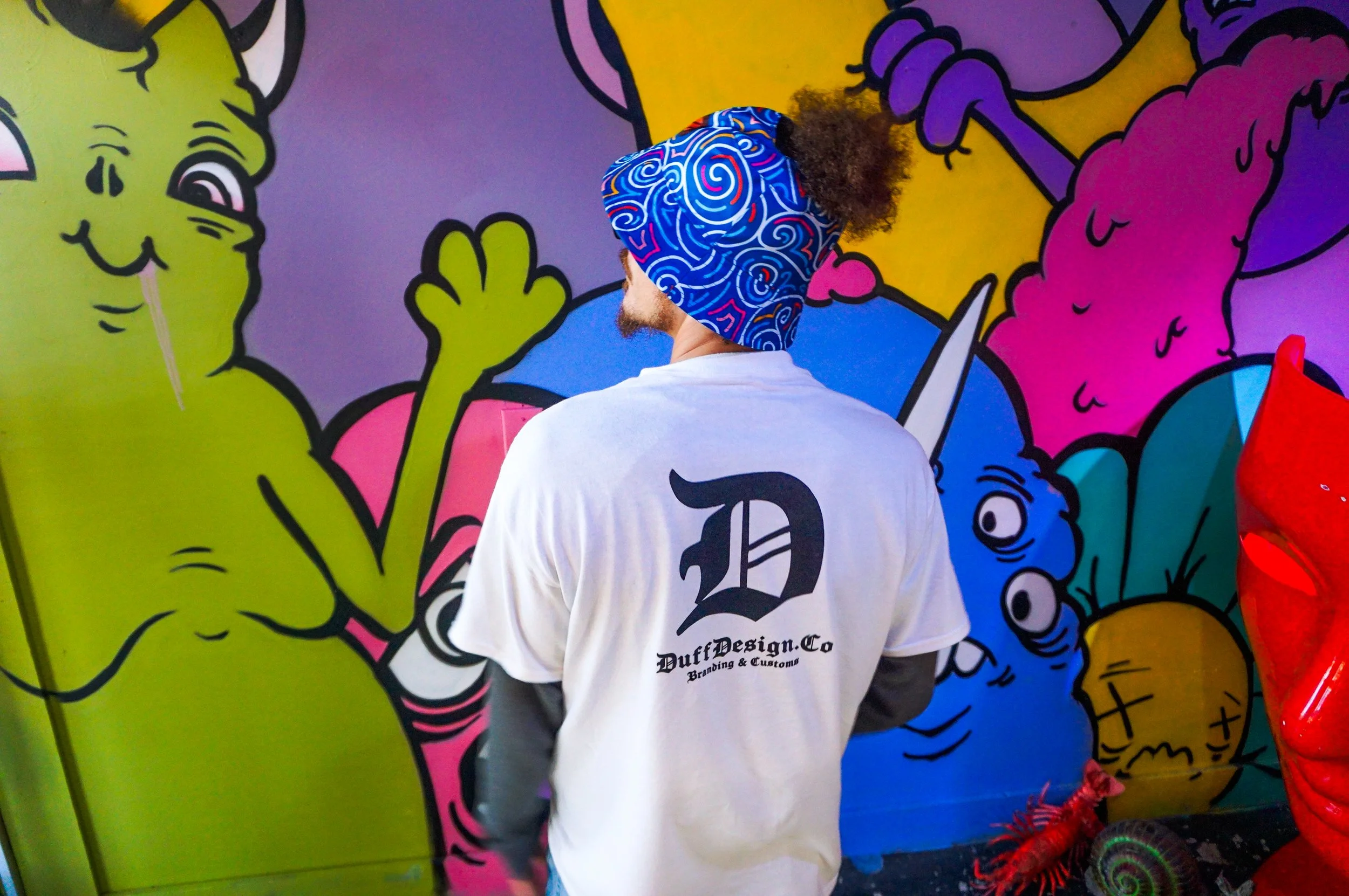 Person with curly hair wearing a modified custom colorful bucket hat and a white t-shirt standing in front of a vibrant graffiti wall in the west coast bodega of connect sand Diego featuring cartoon characters.