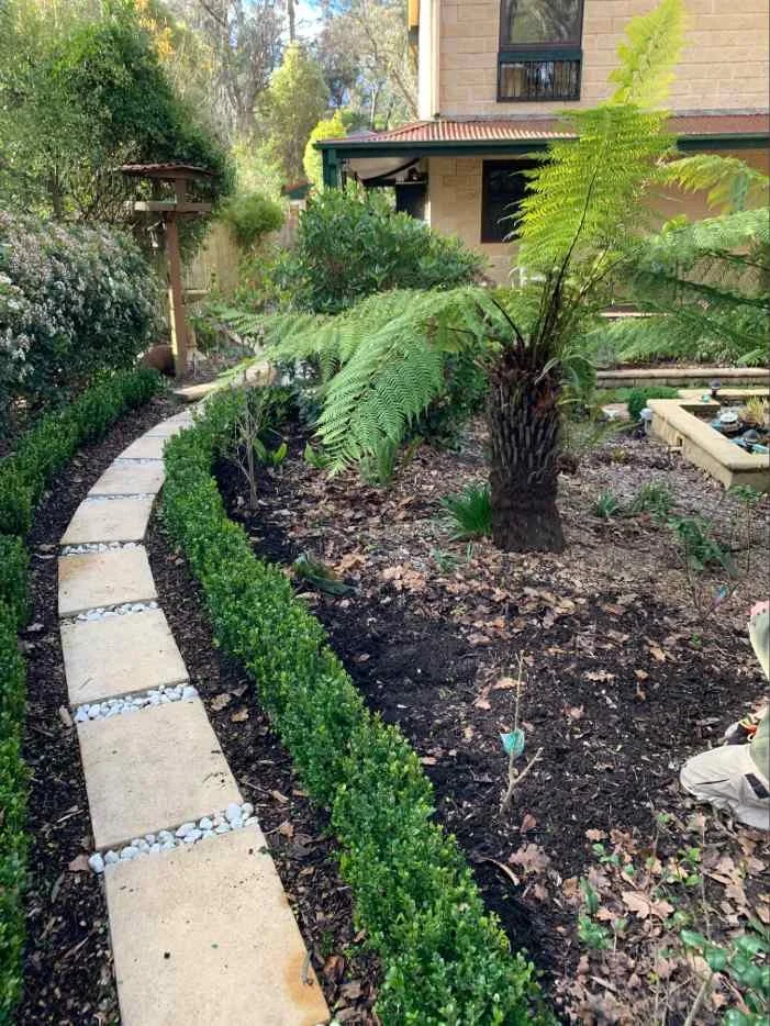 A woman and a man working together in a greenhouse or garden center. The woman is holding a notebook and pen, wearing a straw hat, and smiling. The man is wearing an apron and a backward cap, standing next to a flower bed.