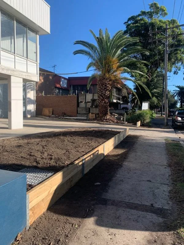A woman and a man working together in a greenhouse or garden center. The woman is holding a notebook and pen, wearing a straw hat, and smiling. The man is wearing an apron and a backward cap, standing next to a flower bed.