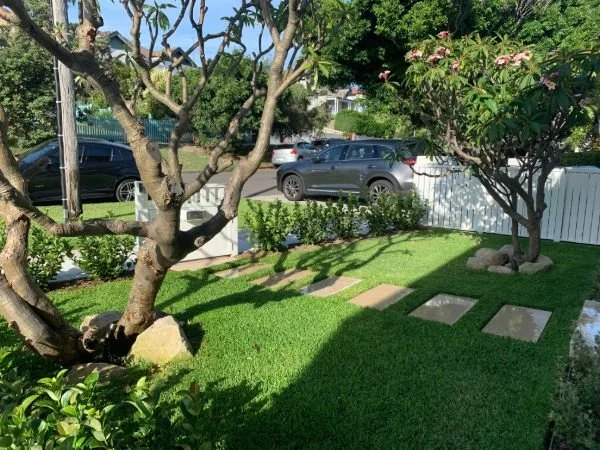 Wooden footbridge over a small waterway in a lush green park with trees and bushes, and a lamp post in the background.