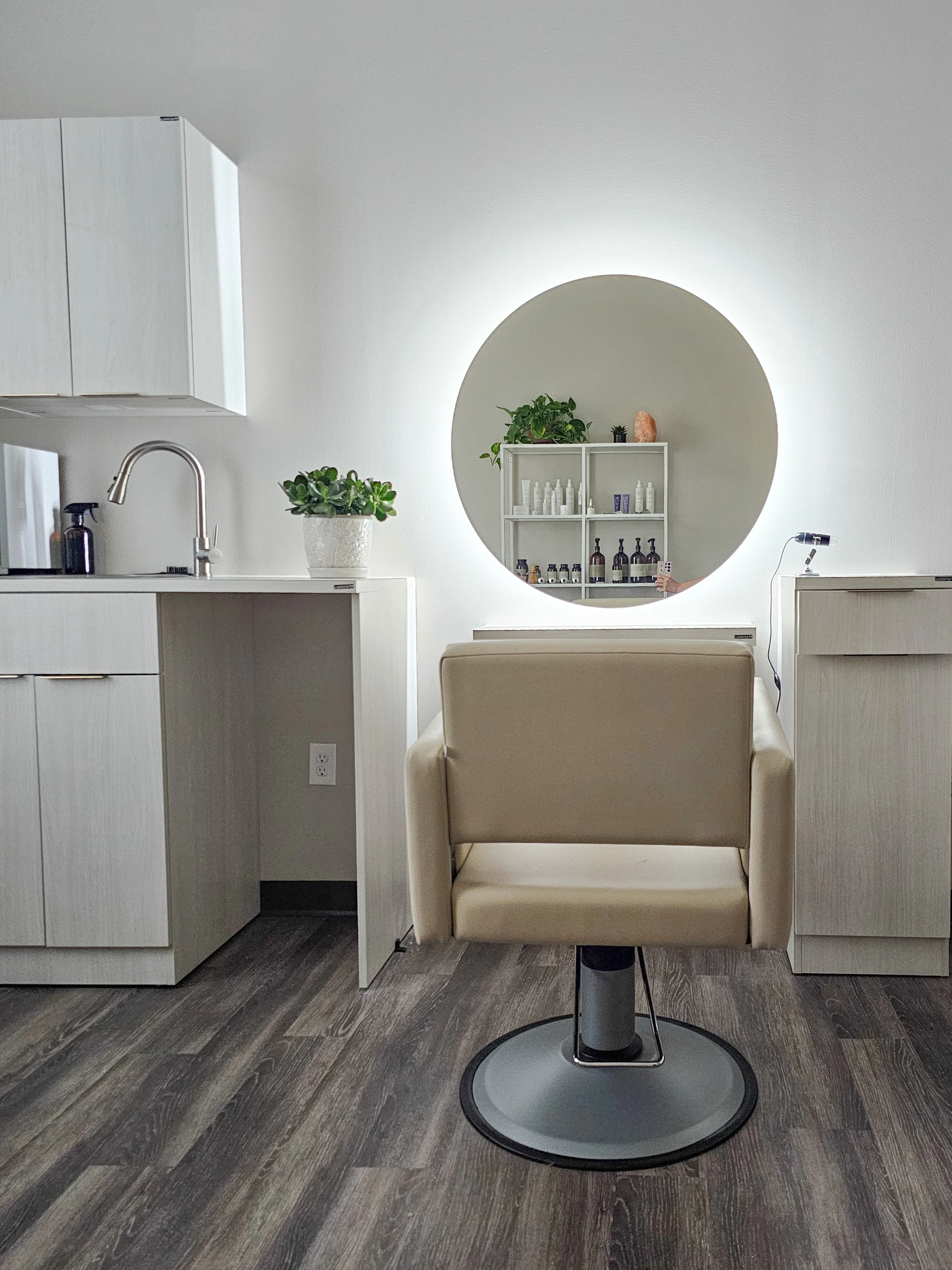 A salon chair in front of a large illuminated round mirror on a white wall. To the left, a white cabinet with a plant on top and a sink with a faucet. To the right, a white cabinet with head spa products.