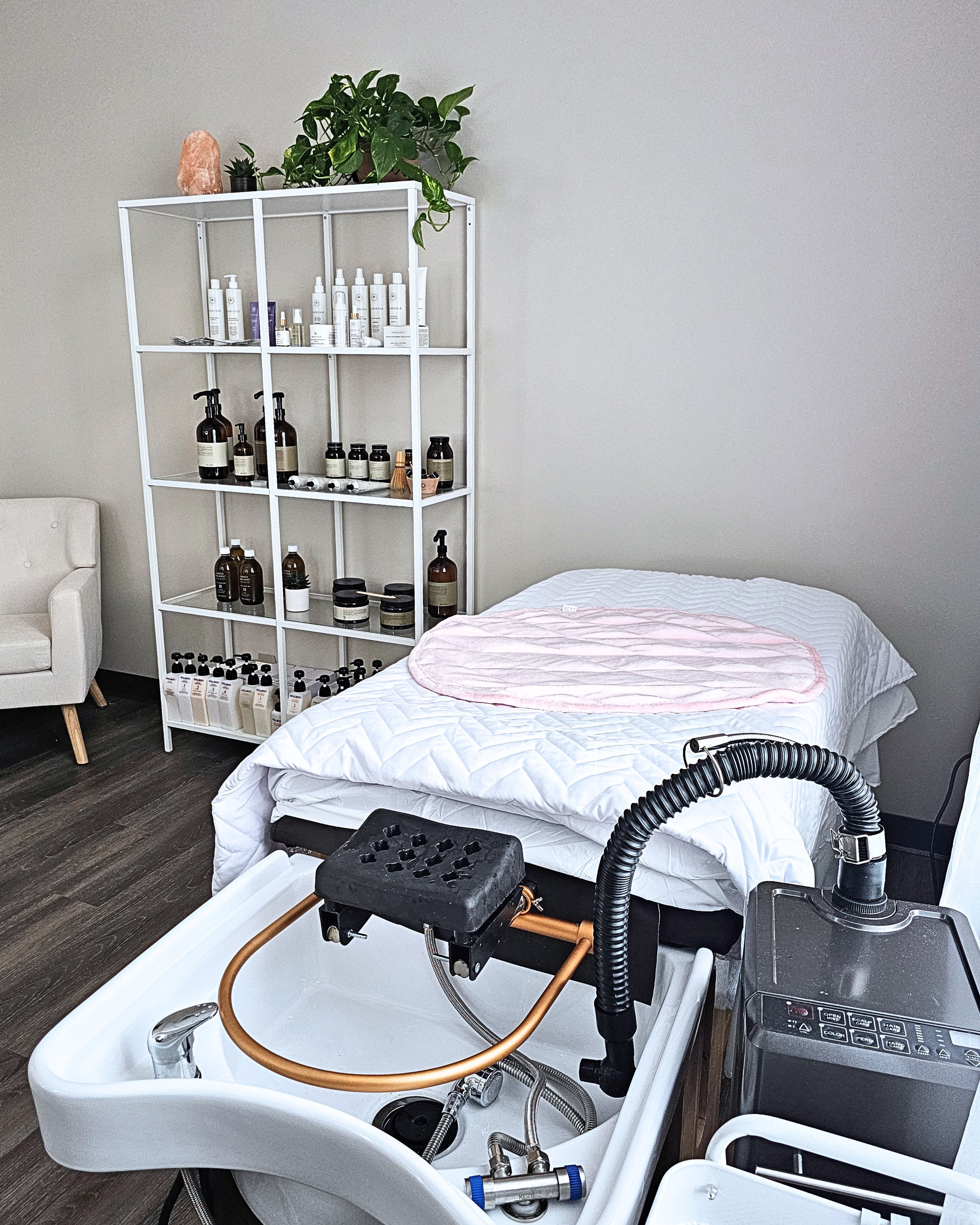 A spa treatment room with a head spa bed, a white shelving unit holding hair care and a basin with equipment, and a small white armchair.