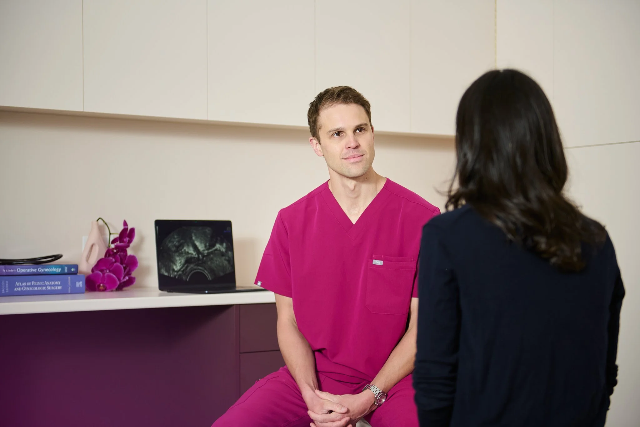 A male doctor in pink scrubs listening to a woman with her back to the camera in a medical office.
