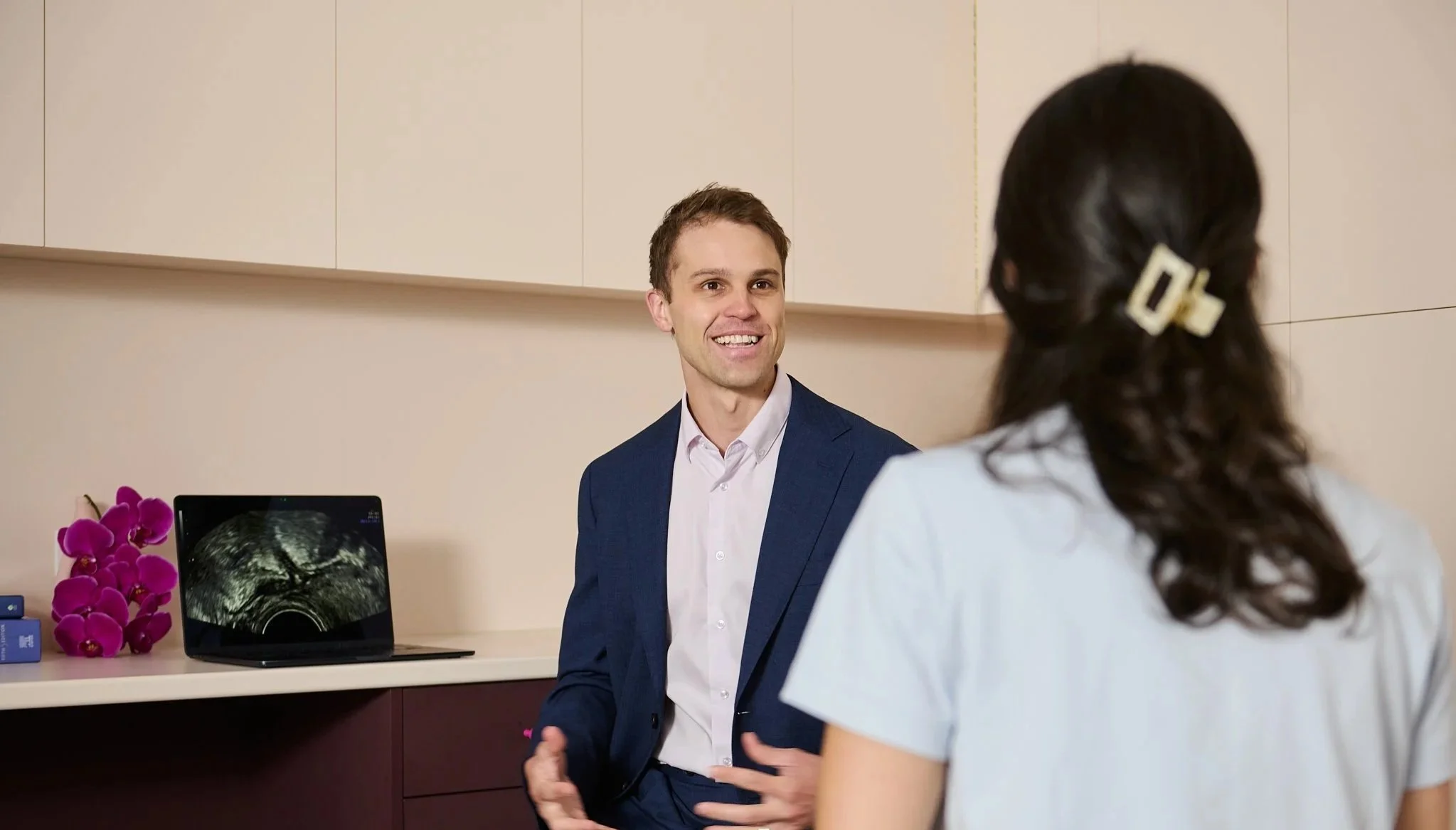 A doctor and a man having a conversation in a medical office, with an ultrasound image on a laptop and a pink orchid on the counter.