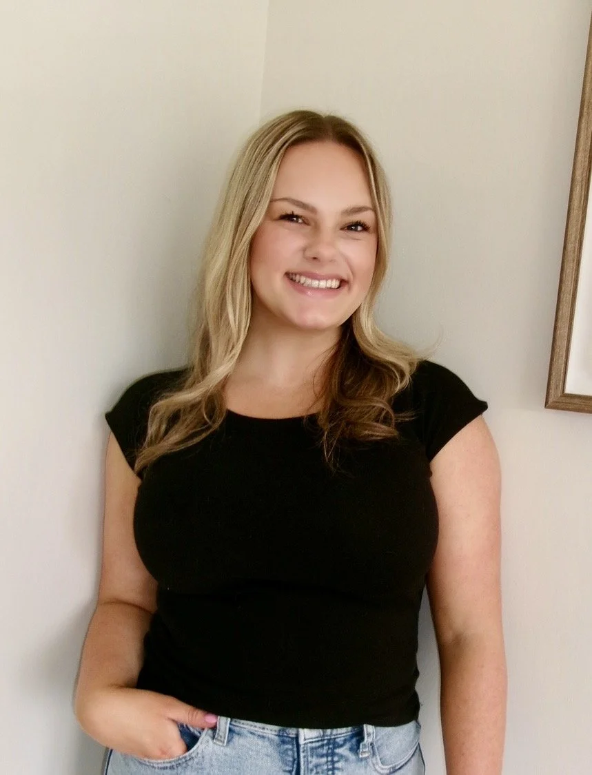A young woman with long, wavy blonde hair, smiling and wearing a black t-shirt and light blue jeans, standing against a plain wall with a picture frame partially visible on the right.