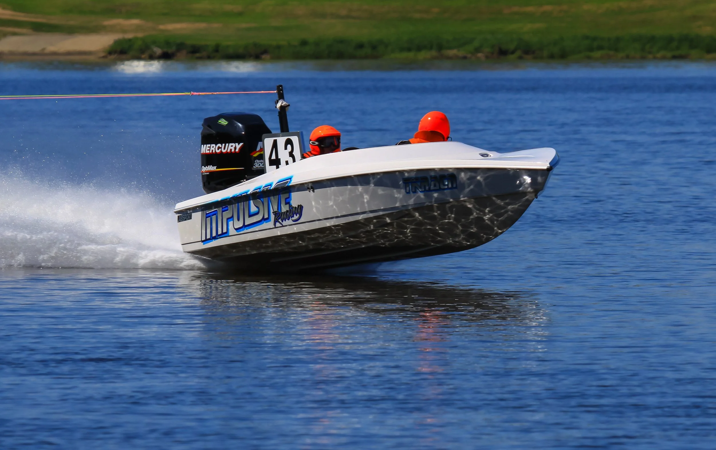 A white racing boat with blue graphics speeding on a body of water, with two people wearing orange helmets onboard and a Mercury engine at the back.