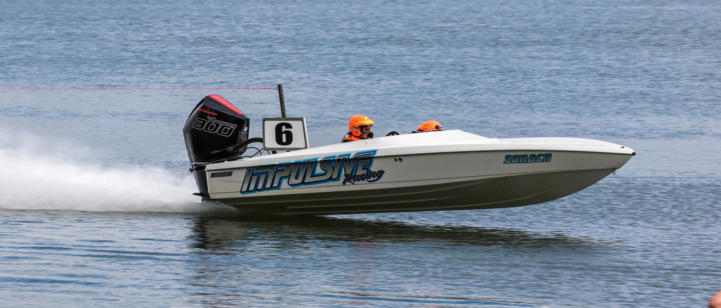 A white speedboat with blue and black decals racing on the water, fueled by a black and red outboard motor, driven by two individuals wearing orange helmets and black goggles.