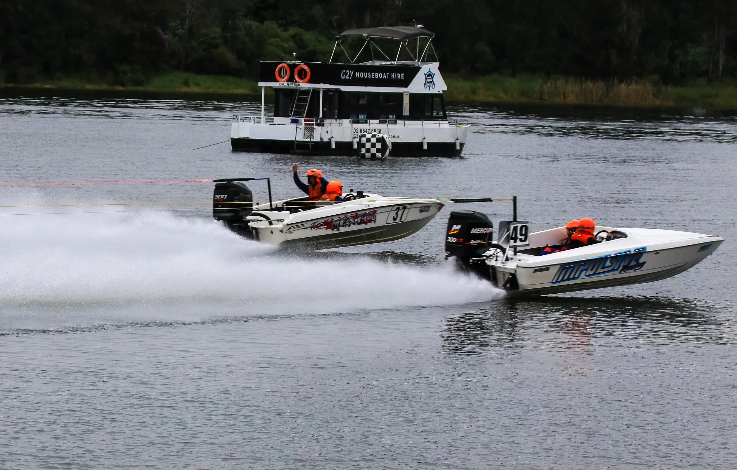 Two speedboats racing on a lake with a houseboat in the background, all wearing orange helmets and advancing at high speed, creating splashes and wakes in the water. speed boat, race boat labsport water ski boat