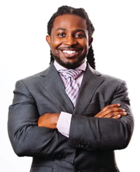 A smiling man in a gray suit with crossed arms, wearing a checkered shirt and tie, standing against a white background.