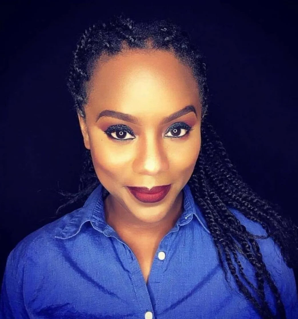Close-up portrait of a woman with braided hair, wearing a blue shirt and makeup with bold lipstick and eye makeup, against a black background.