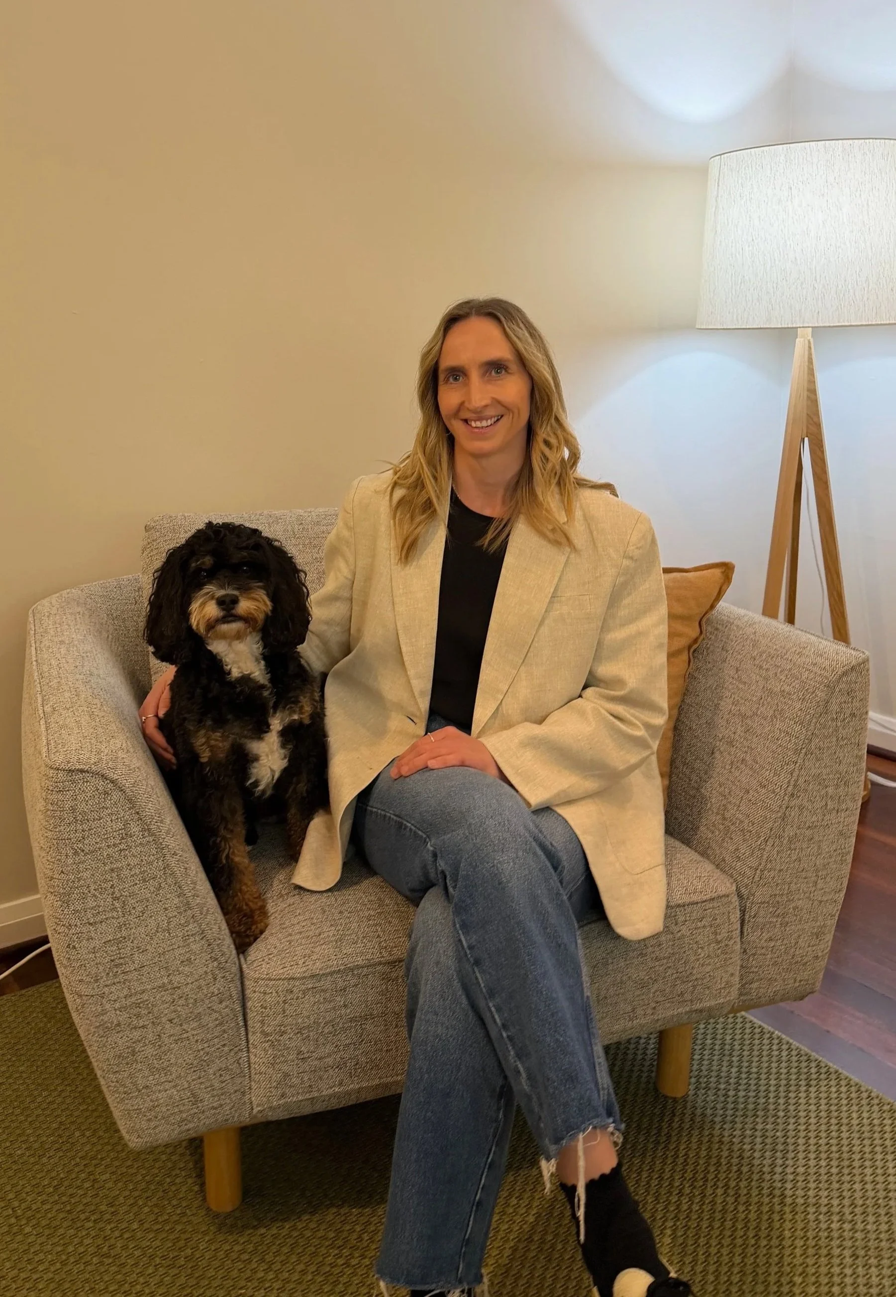 A woman with wavy blonde hair sitting on a beige armchair with a small black and brown curly-haired dog beside her. She is smiling, wearing a cream blazer, black shirt, blue jeans, and black sneakers, in a cozy living room with a tall floor lamp and pillows.