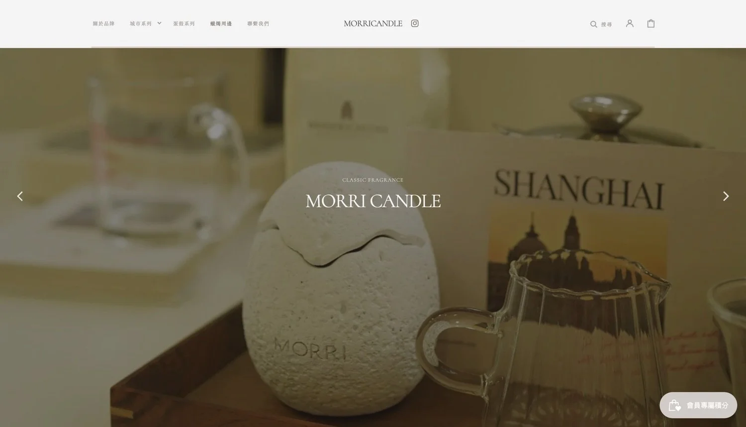 A textured white candle with the word 'MORRI' engraved on it, sitting on a wooden tray, with a glass measuring cup nearby, and a box labeled 'SHANGHAI' in the background.