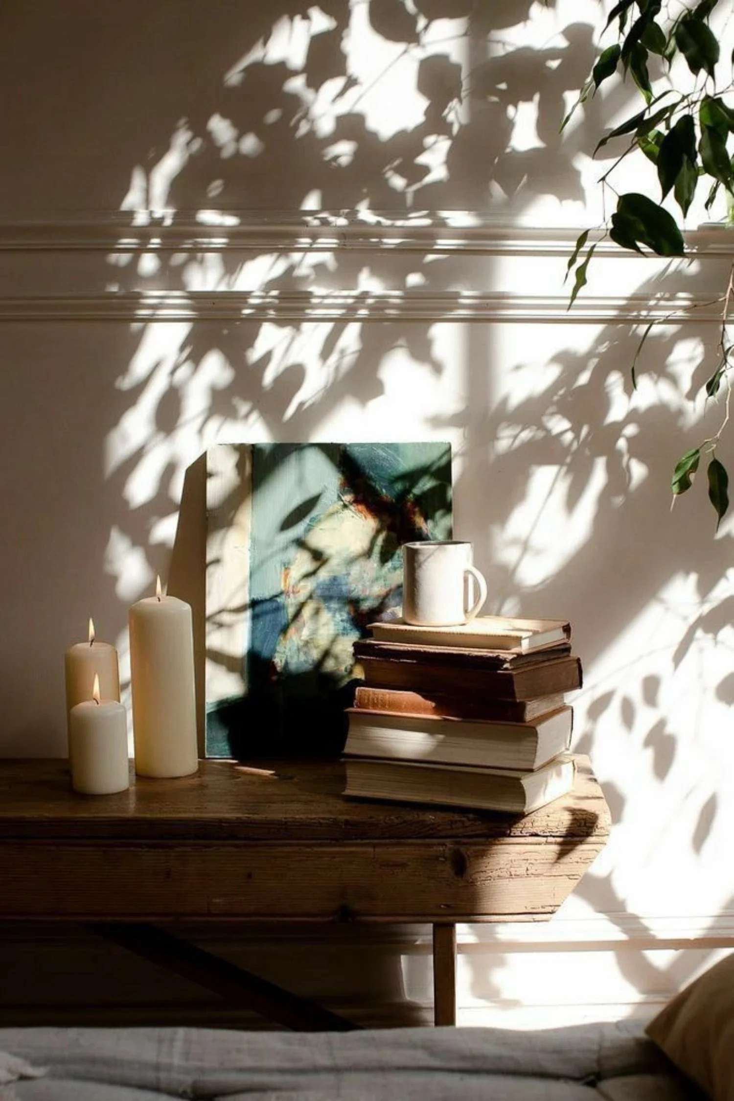 Sunlight creates shadow patterns of leaves on a white wall above a wooden table with stacked books, a white mug, and three lit candles, with a green leafy plant hanging nearby.