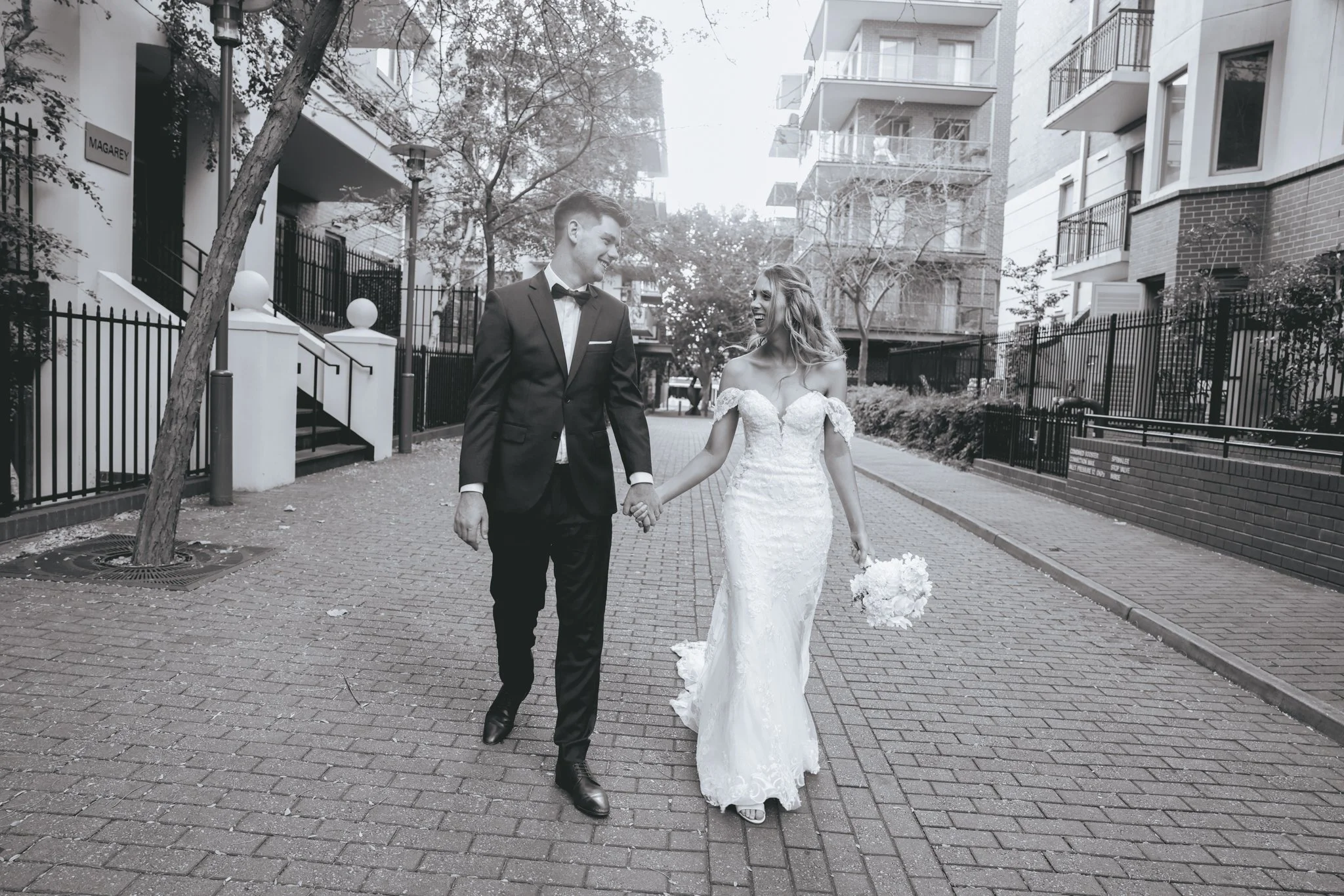Bride and groom walking outdoors on a paved path, holding hands and smiling at each other, with modern apartment buildings and trees in the background.