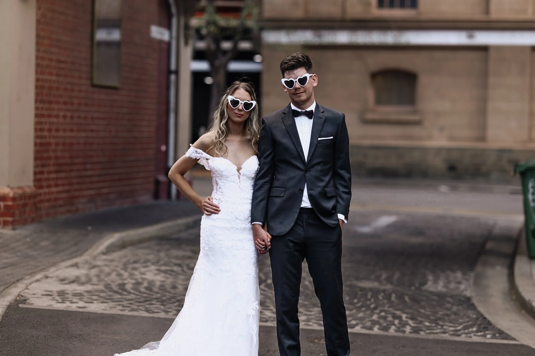 A newly married couple in wedding attire wearing heart-shaped sunglasses, standing hand in hand outdoors on a cobblestone street, with brick and beige building backgrounds.