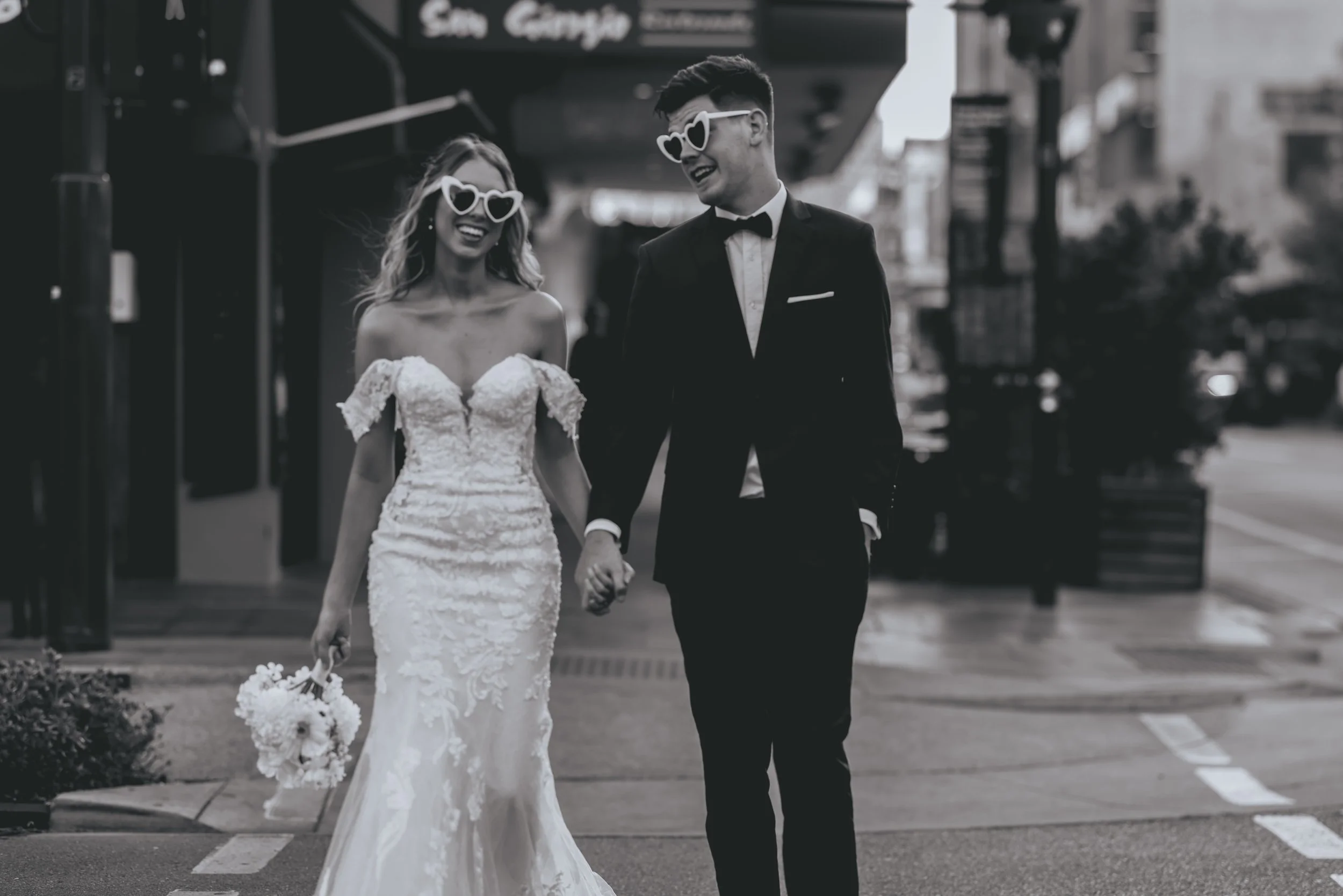 A bride and groom holding hands and smiling while walking on a city sidewalk. The bride is wearing a lace wedding gown and holding a bouquet, and both are wearing heart-shaped sunglasses.