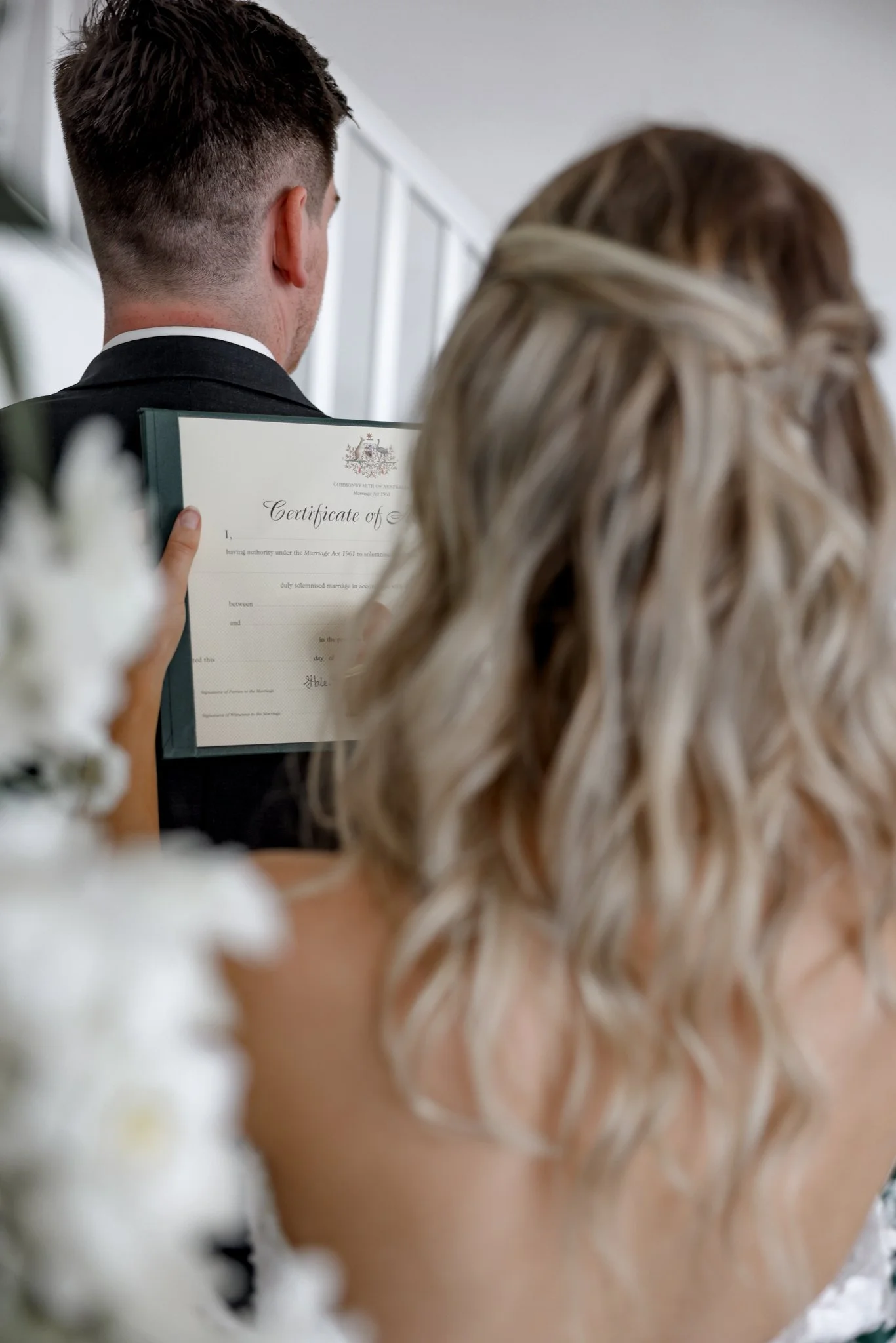 A bride and groom at their wedding ceremony, with the groom holding a certificate of marriage, seen from behind.
