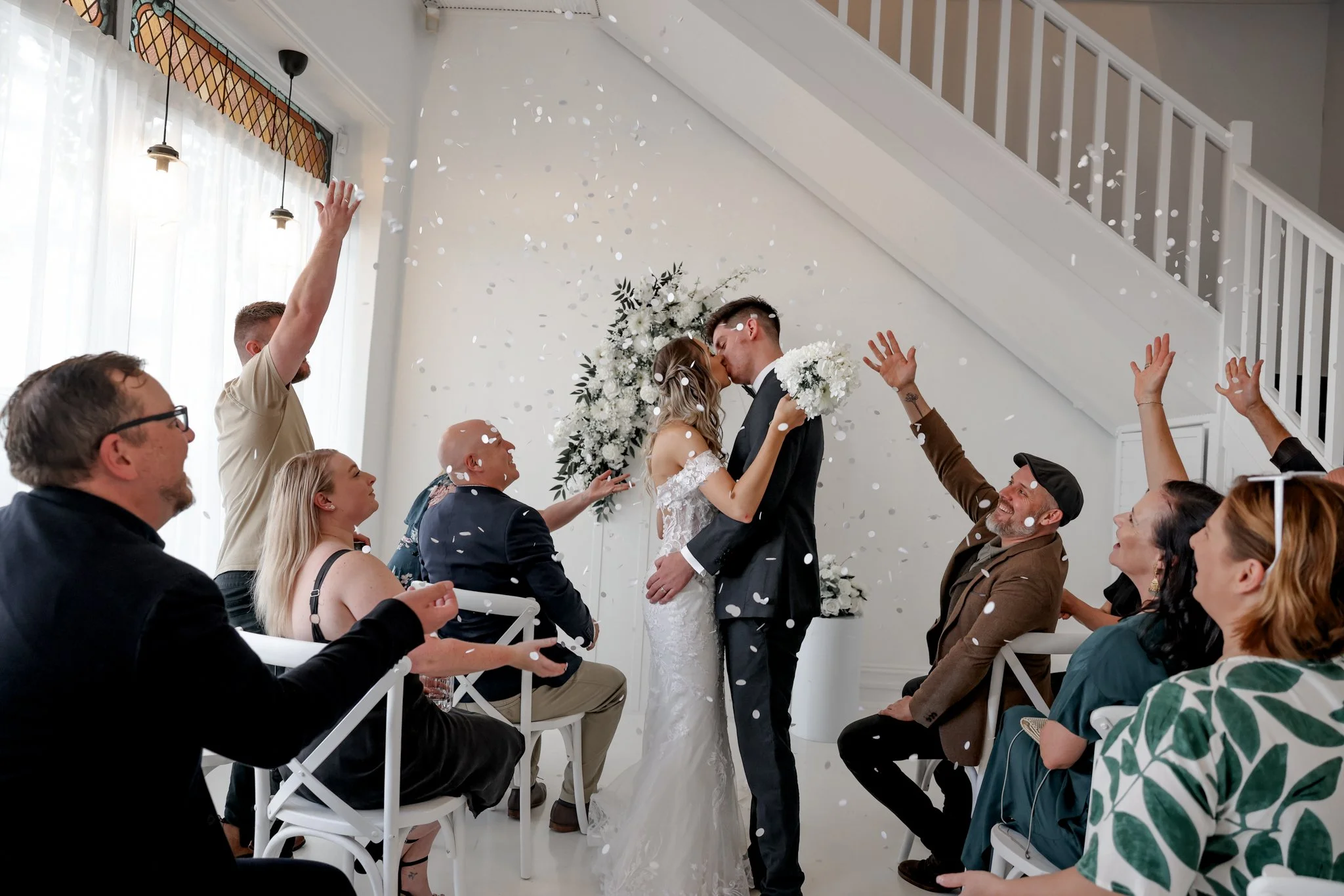 A bride and groom kiss at their wedding ceremony surrounded by seated guests throwing white confetti, with a white floral arrangement behind them in a bright room with large windows.
