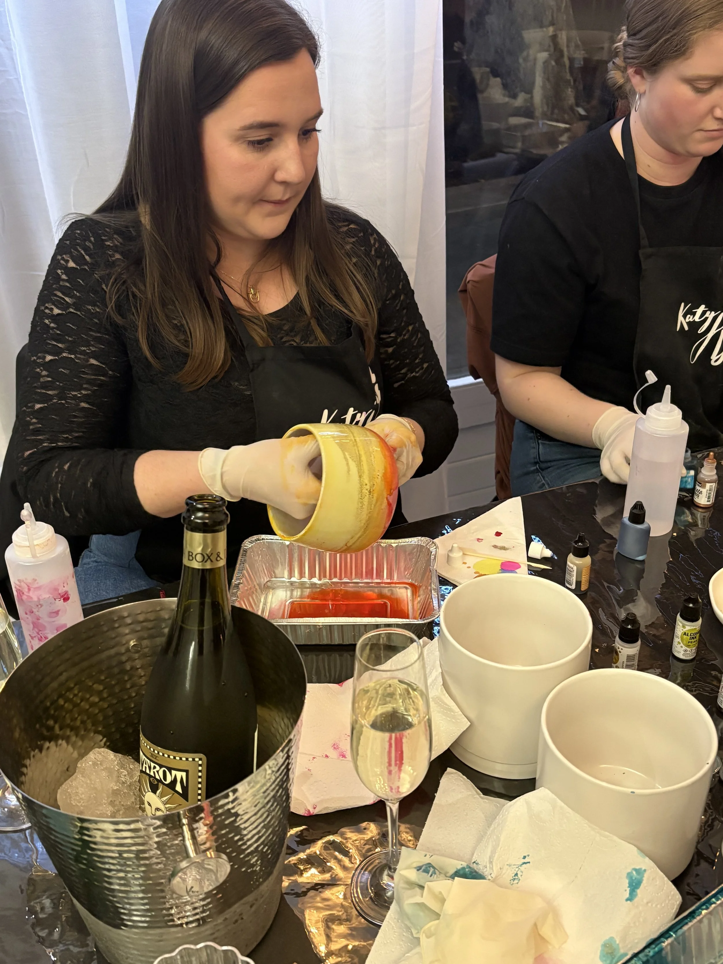 Two women are pouring paint or similar liquid from a container into another container at a worktable, with supplies like bottles, cups, and paper towels around them.