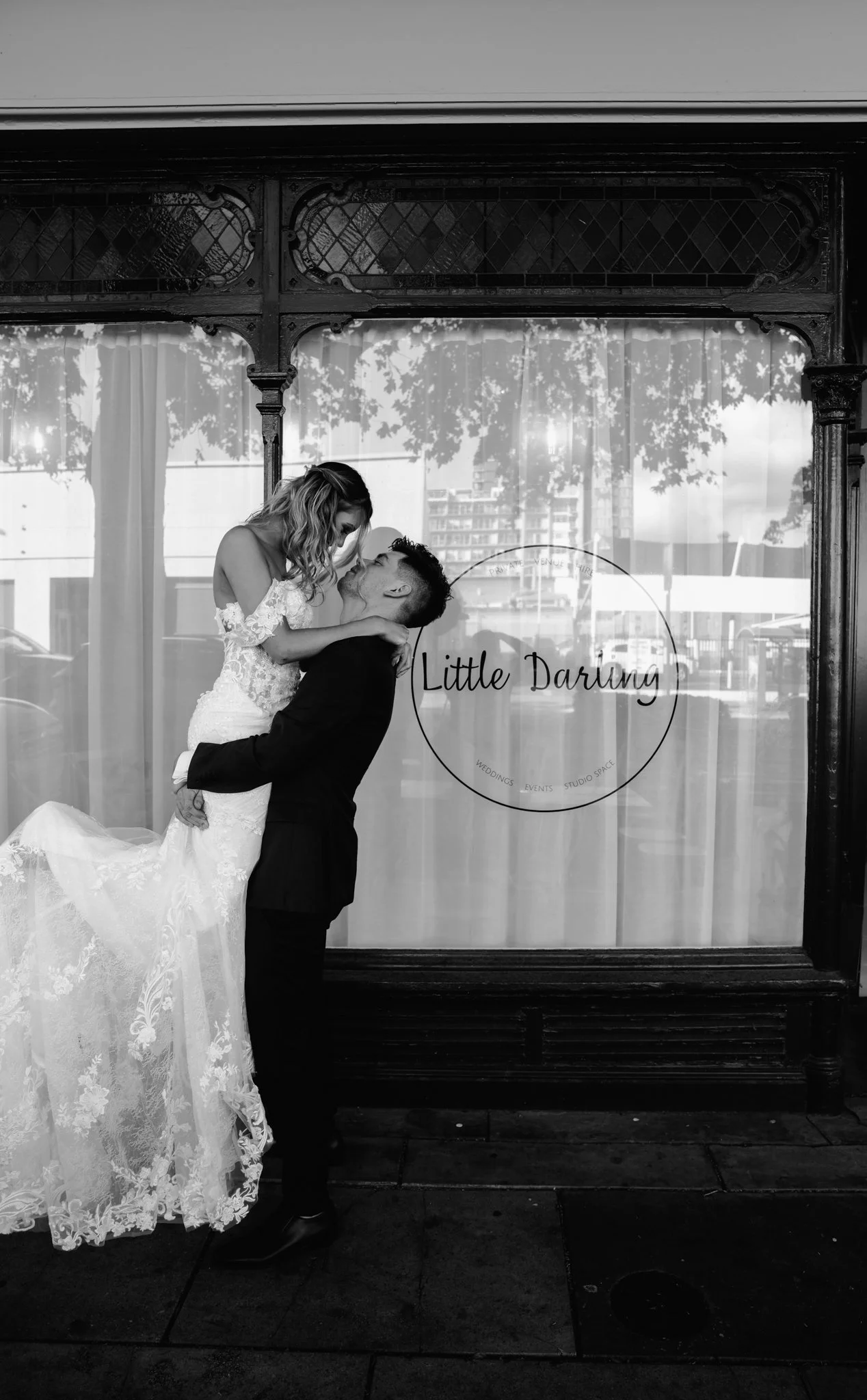 A black-and-white photo of a couple on their wedding day, with the groom lifting the bride. They are in front of a store window with the sign 'Little Darling'.