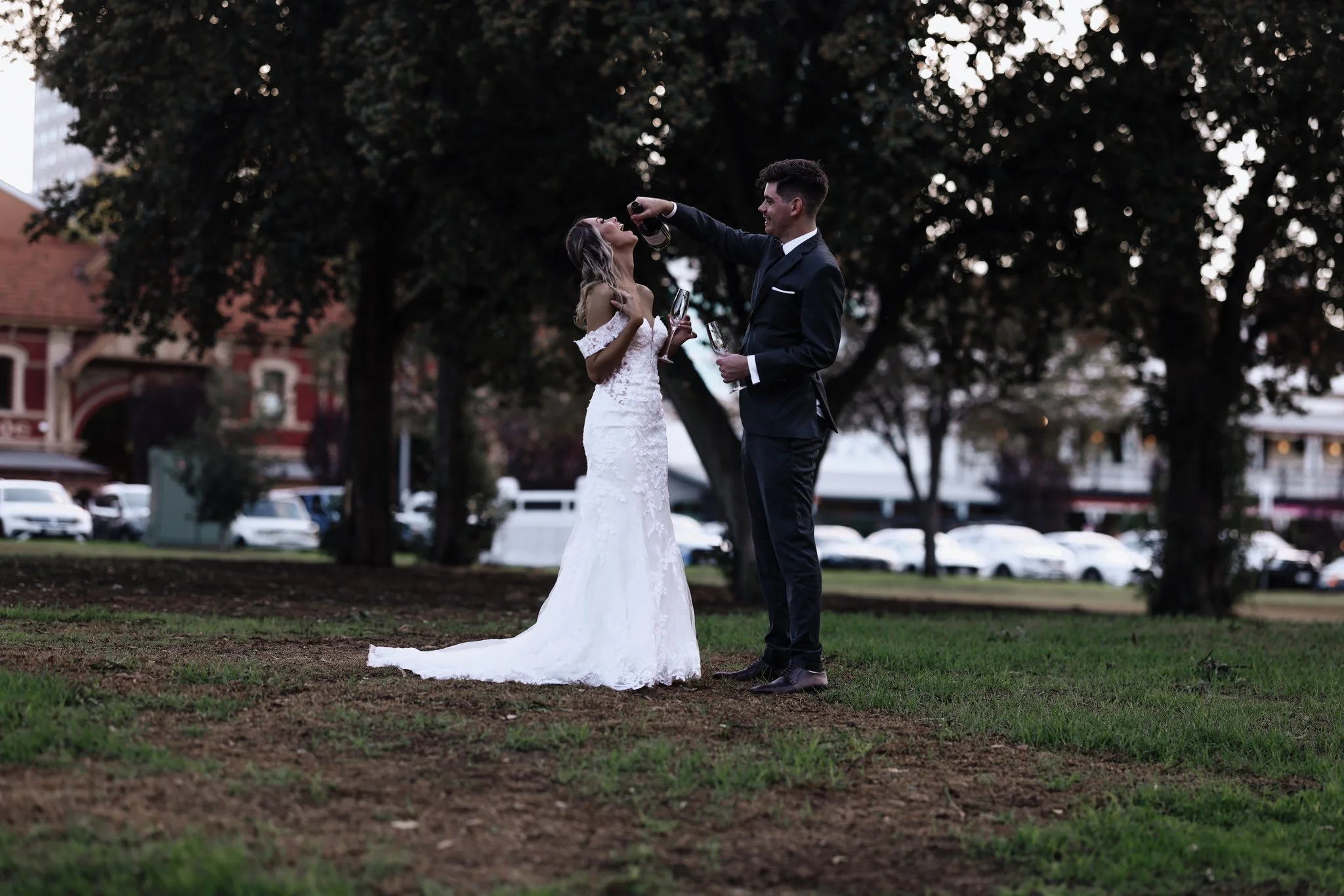 A bride and groom celebrating their wedding outdoors, with the groom pouring champagne into the bride's mouth, standing on grass with trees and parked cars in the background.