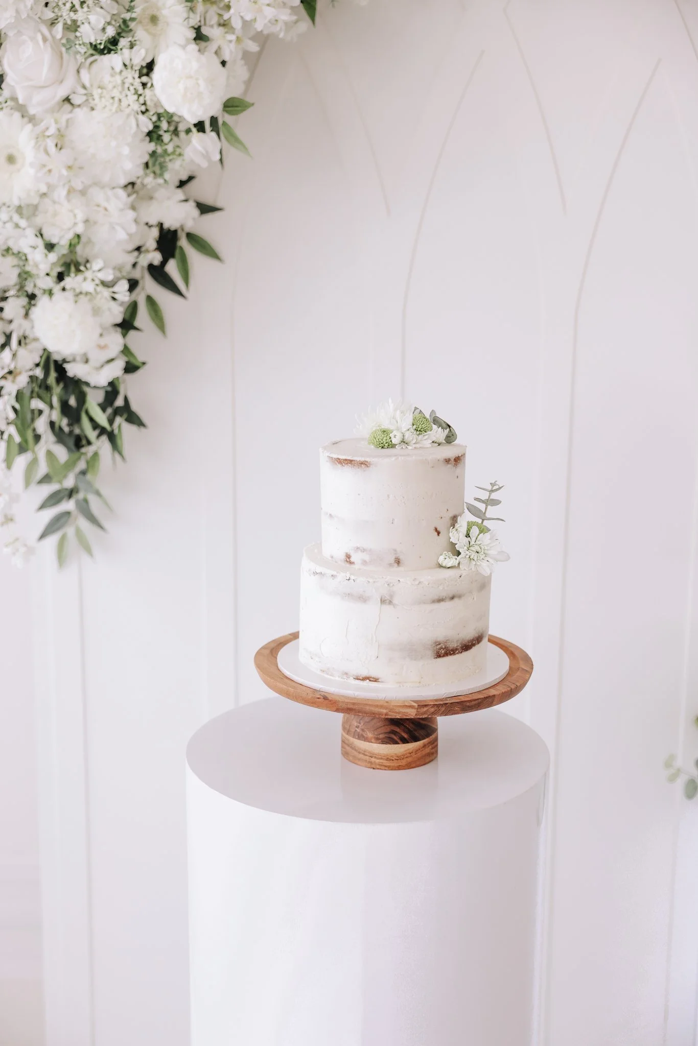 A two-tiered white wedding cake with minimal frosting, decorated with small white flowers and greenery on top and on the side, placed on a wooden cake stand on a white cylindrical pedestal against a white wall with floral arrangements.